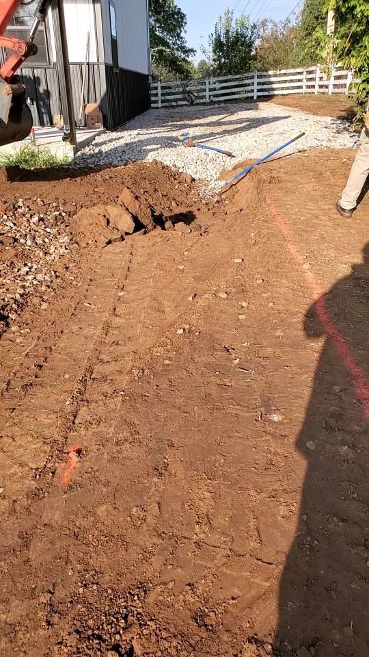 A construction scene showing an excavator arm, a pile of loose soil, and gravel paving in a yard by a white fence.