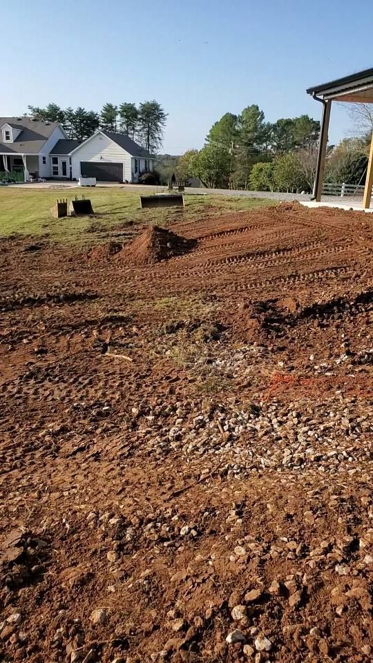 A view of a construction site with exposed red dirt, tire tracks, and nearby residential houses under a clear blue sky.