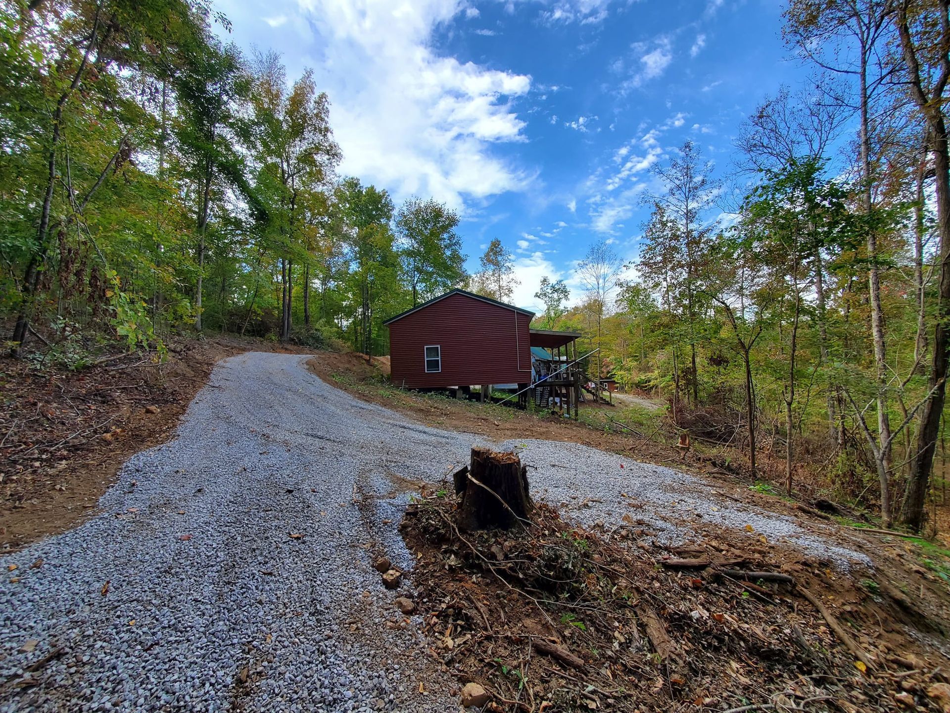 A red cabin sits at the end of a long, freshly laid gravel driveway surrounded by trees under a bright blue sky.