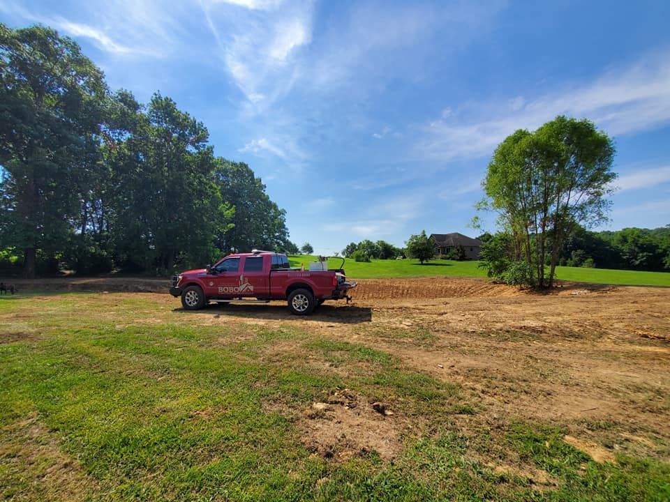 A red pickup truck parked on a cleared dirt lot in a rural setting with trees and a bright blue sky.