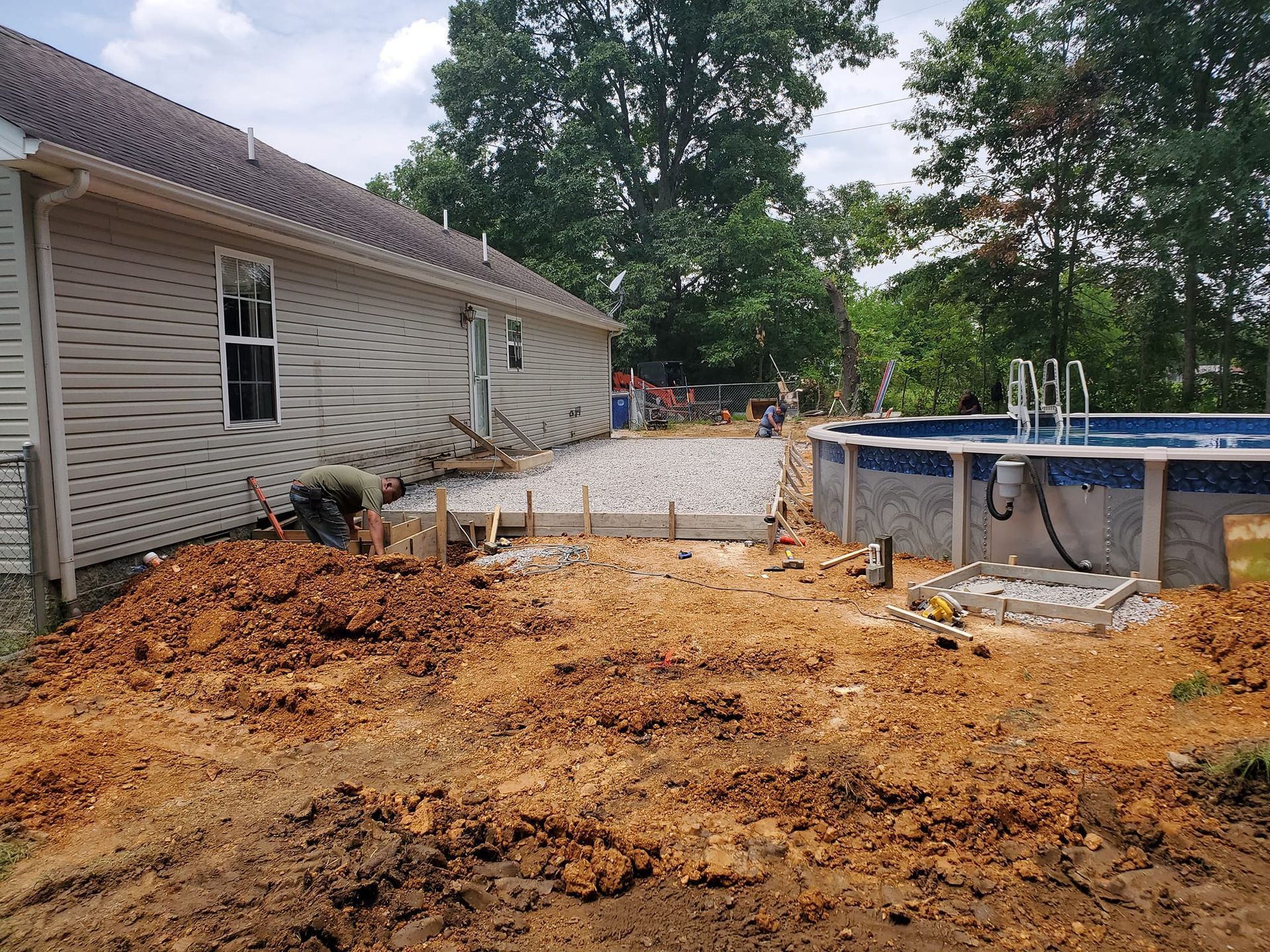 A person works near a house on a concrete patio foundation being prepared next to an above-ground swimming pool.