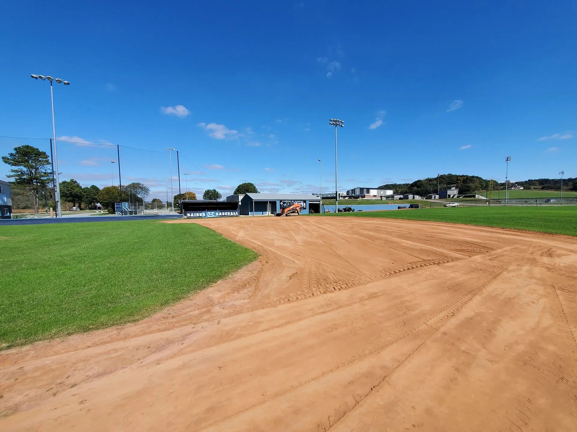 A dirt baseball infield surface under a clear blue sky, bordered by bright green grass and stadium light poles.