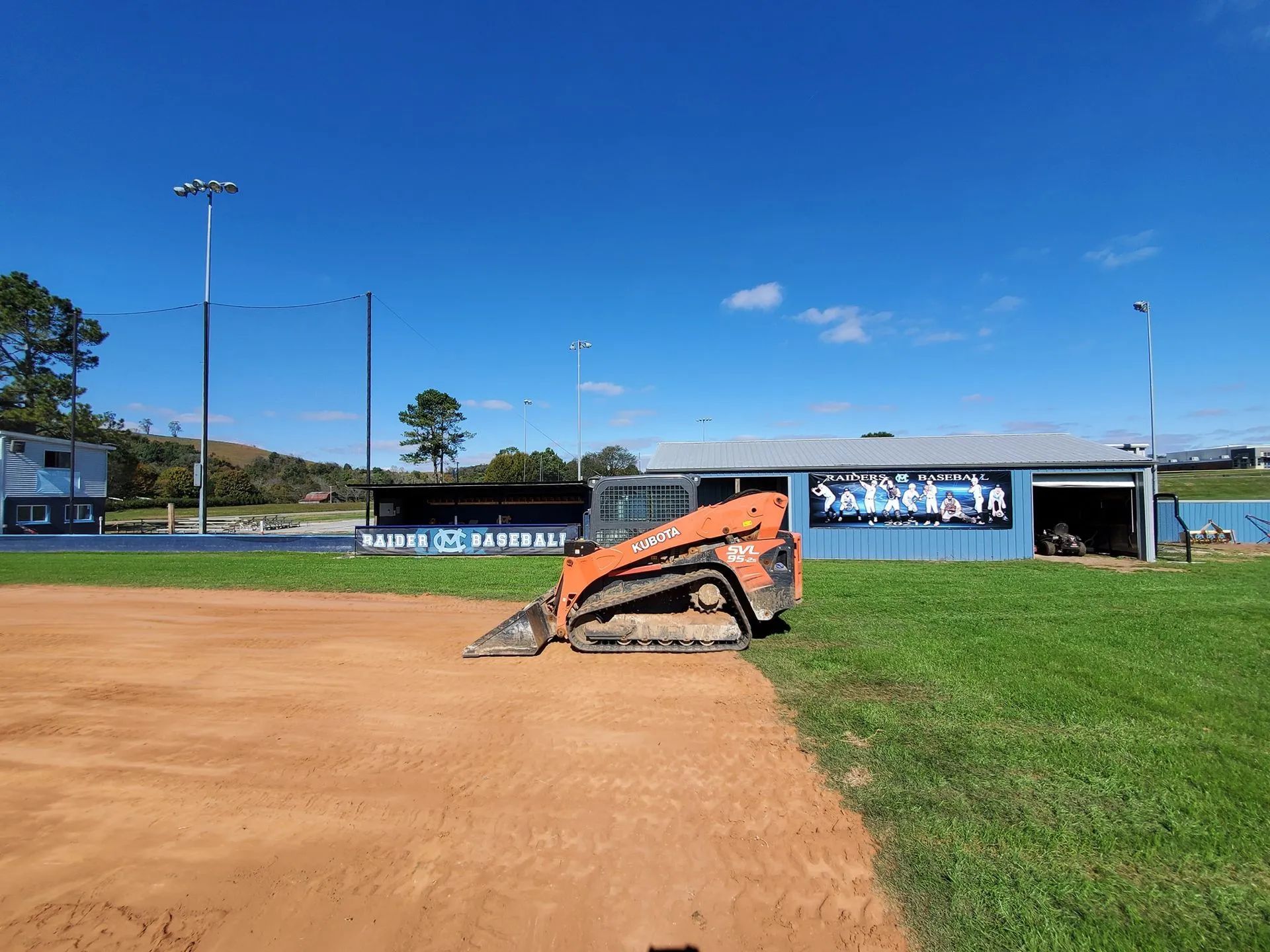 An orange tracked skid steer sits on a dirt baseball infield next to a green grass field and a blue equipment building.