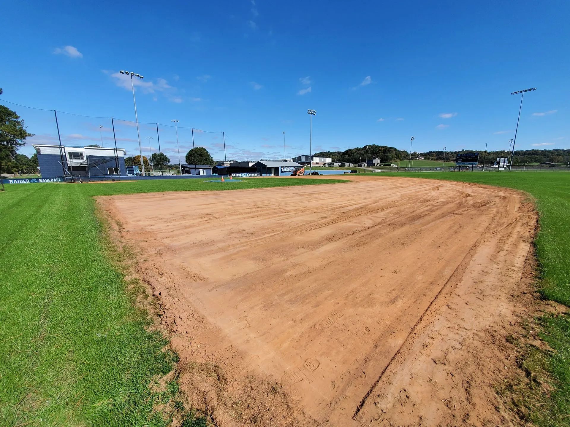 A large, raked dirt infield of a baseball or softball diamond sits amidst green grass under a clear blue sky.