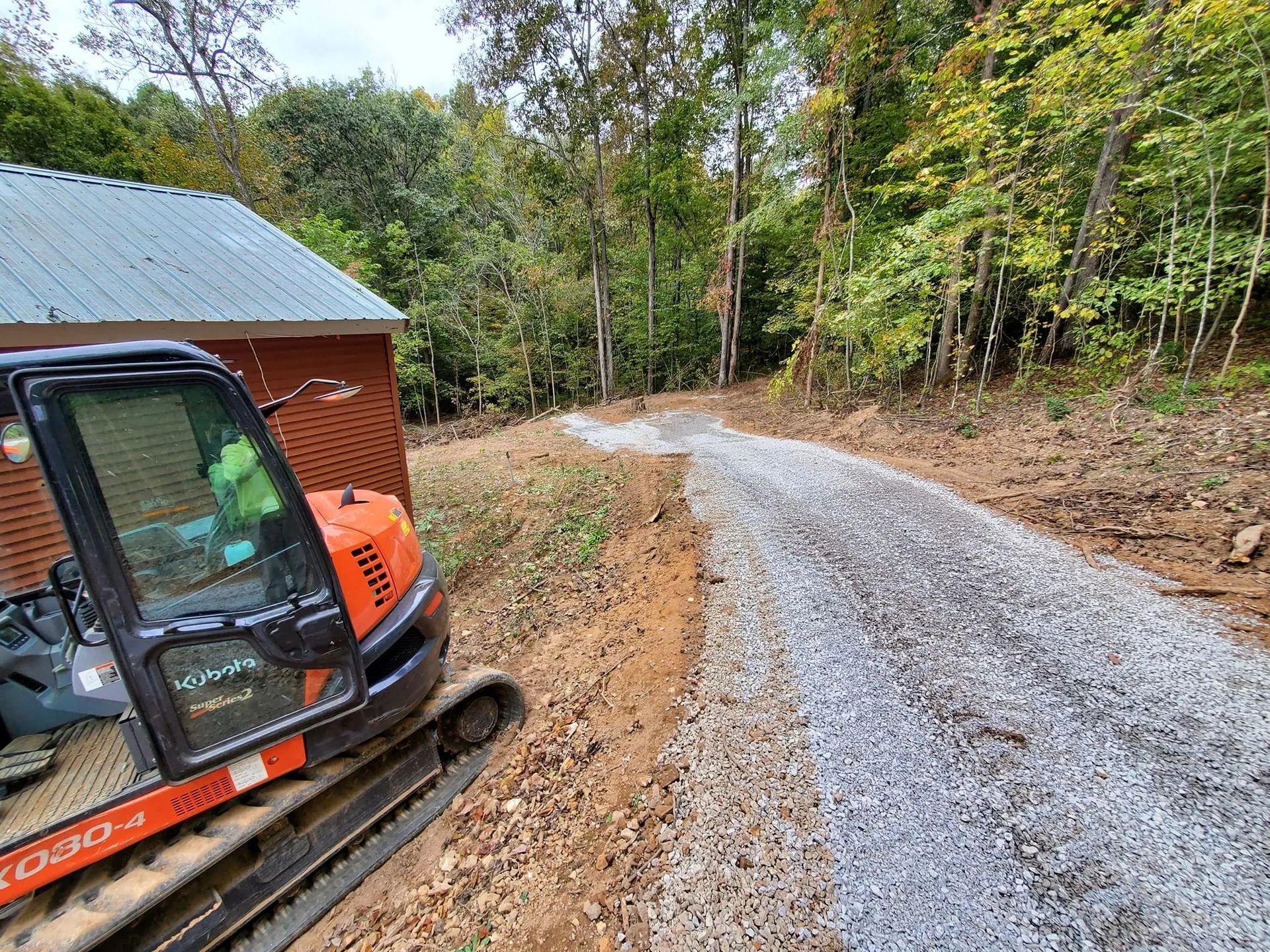 An orange excavator sits beside a newly laid gravel path leading into a wooded area next to a red shed.