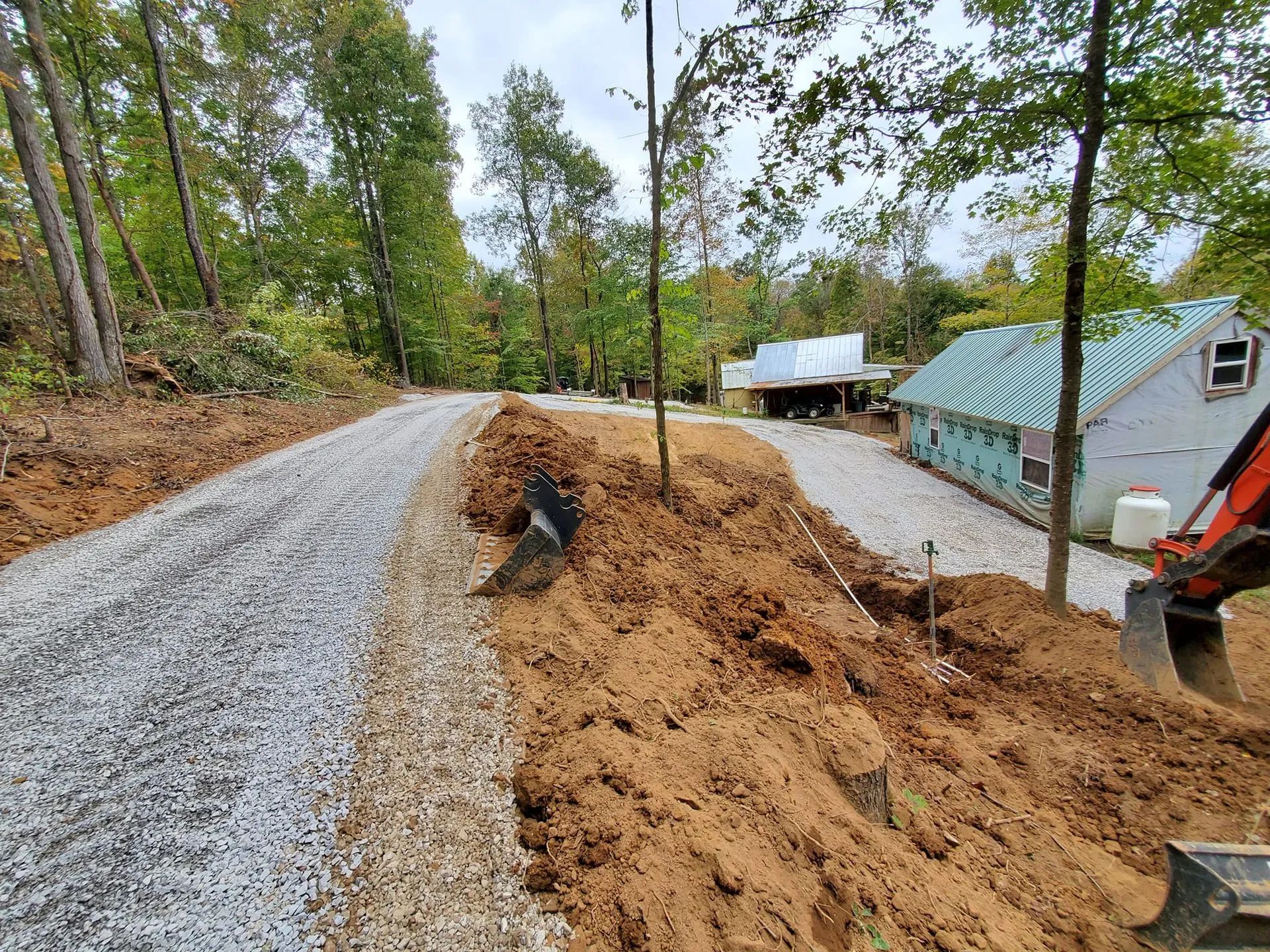 A dirt and gravel driveway under construction, with a large pile of excavated soil and heavy machinery nearby.