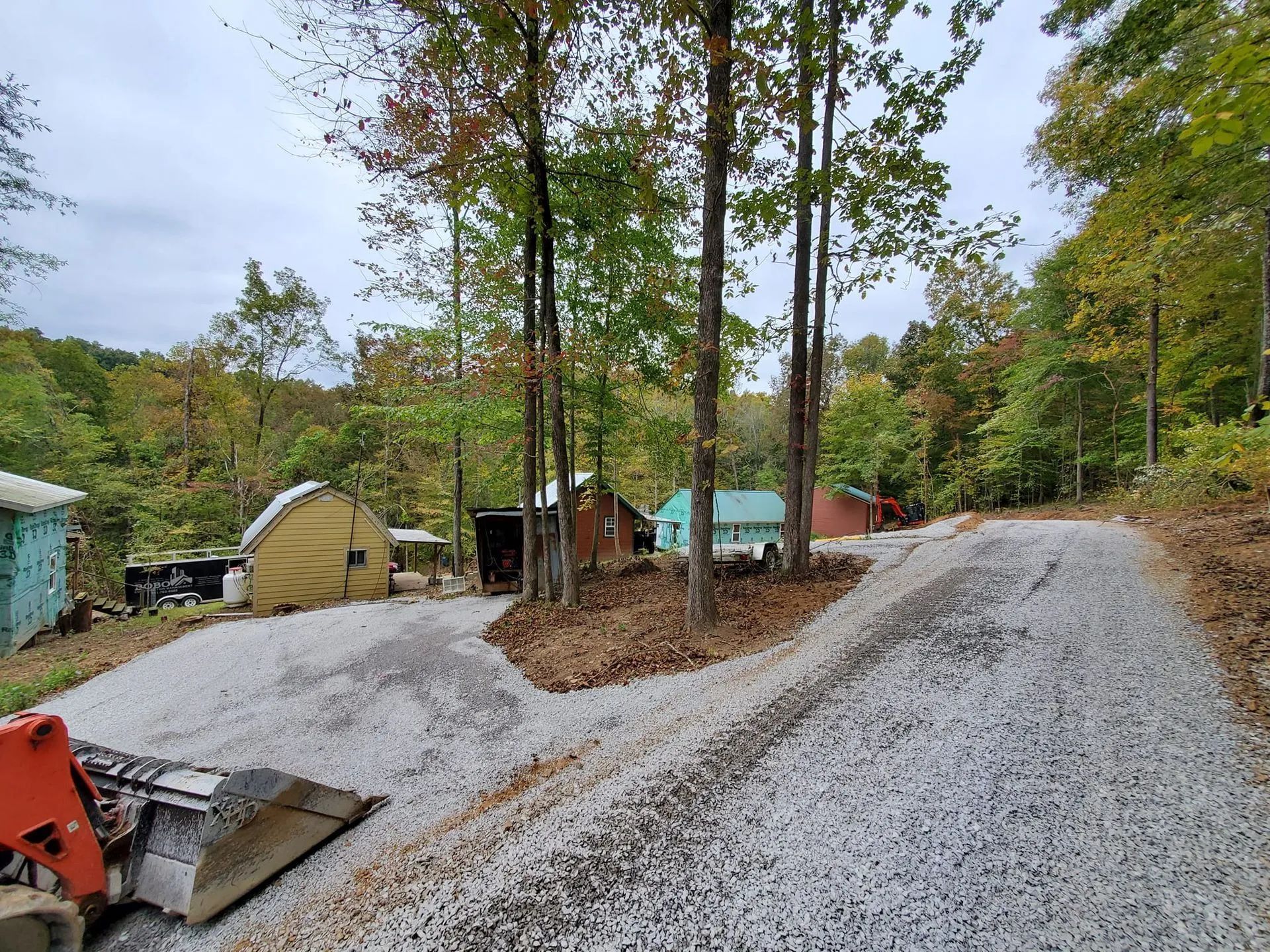A front loader sits on a gravel drive leading toward several small cabins nestled among trees on an autumn hillside.