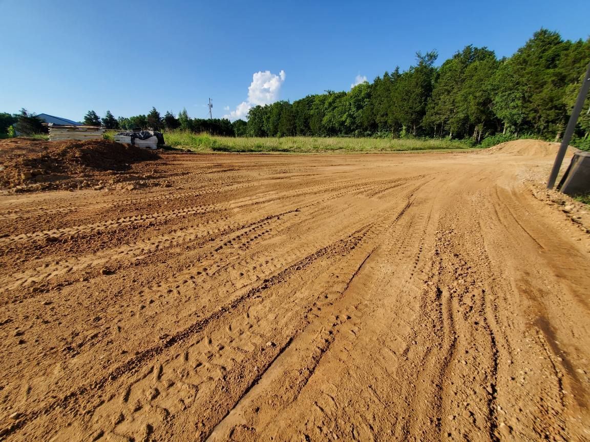 A construction site featuring a cleared, dirt-covered lot with tire tracks, bordered by a line of green trees under blue sky.