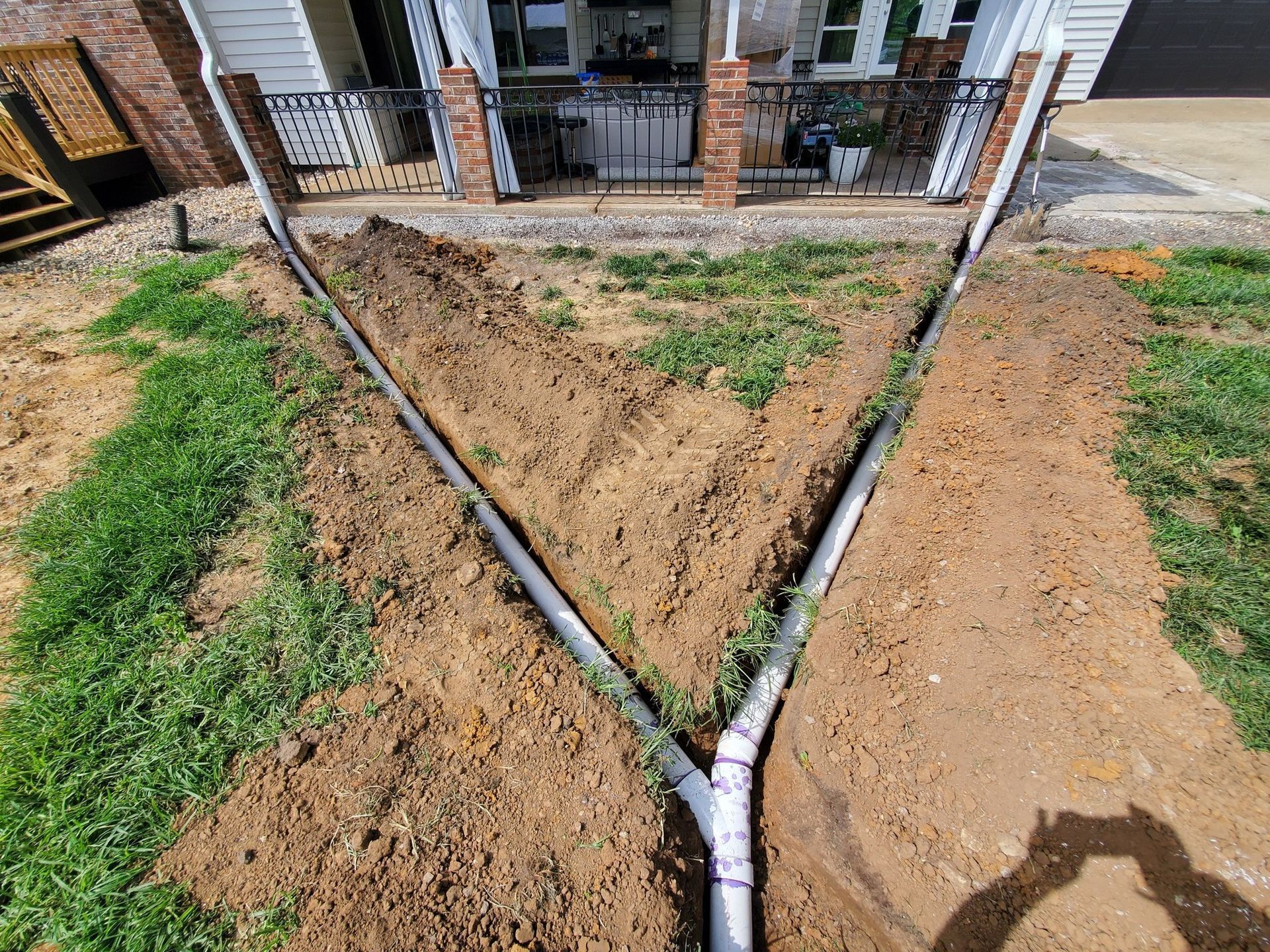 A Y-shaped PVC pipe drainage system installed in a residential yard trench leading away from a house foundation.