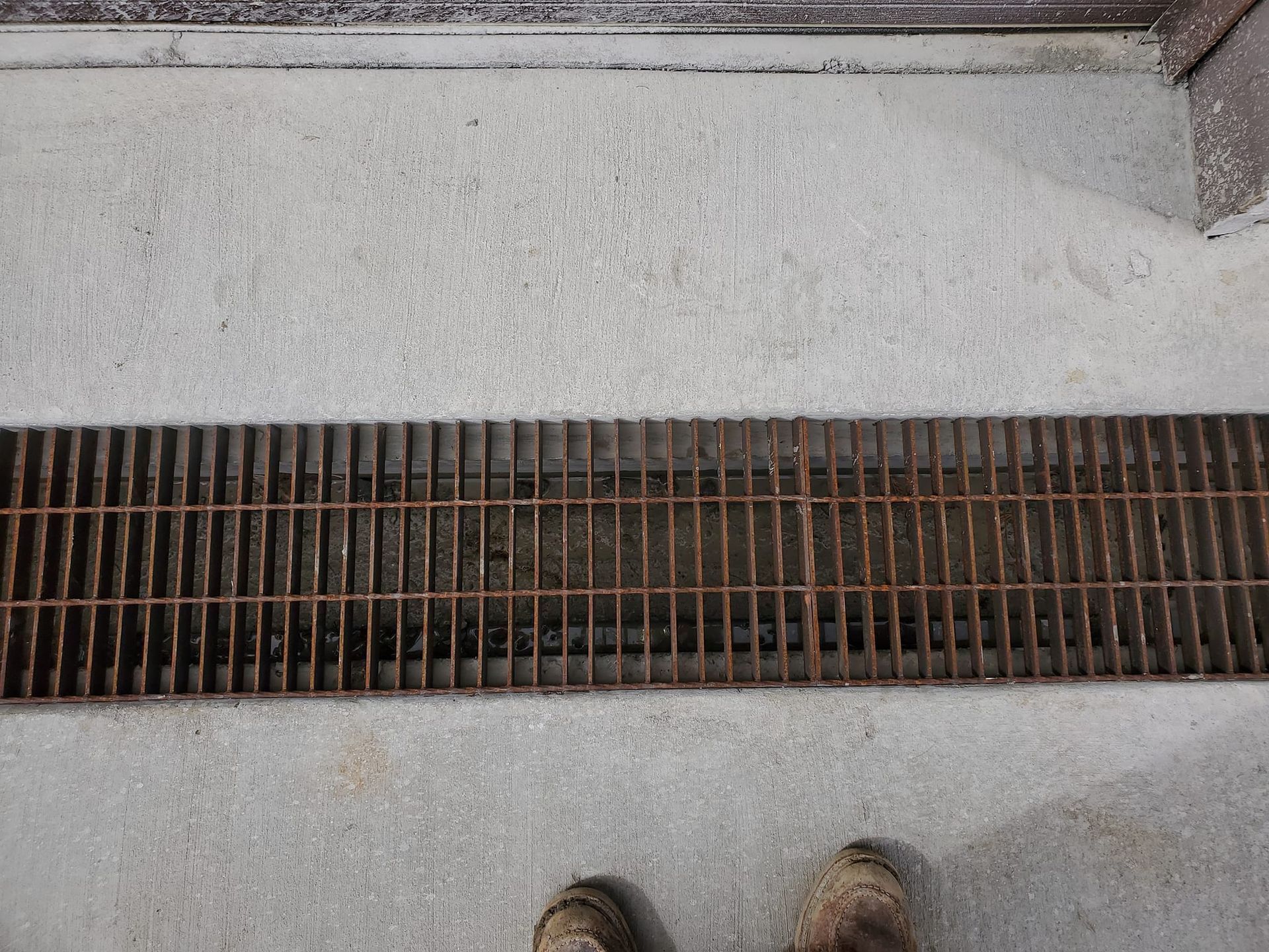A metal trench drain grate embedded in a concrete walkway, viewed from above with a person's boots visible at the bottom.
