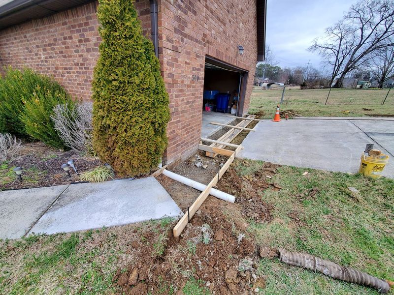 Construction forms for a new concrete walkway are set up next to a brick house garage, with a white pipe placed underneath.