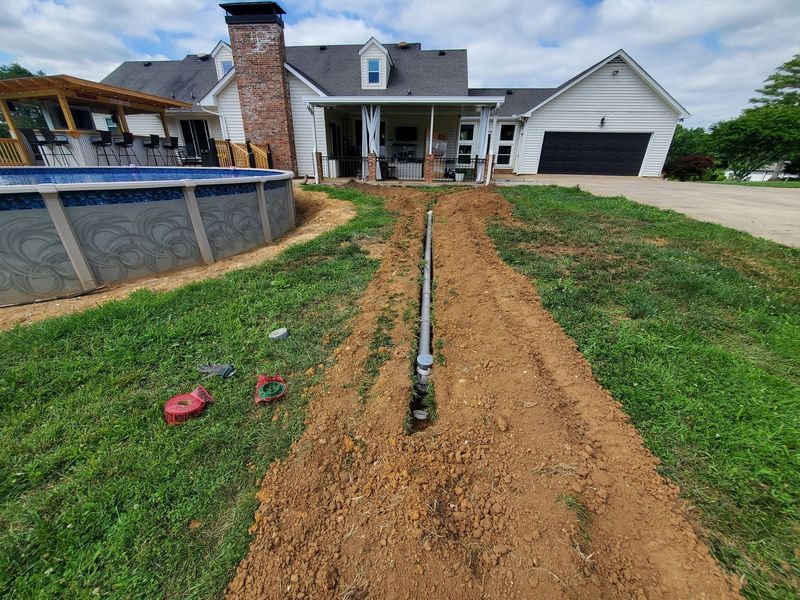 A trench with a pipe running through a grass yard toward a house with a pool in the background.