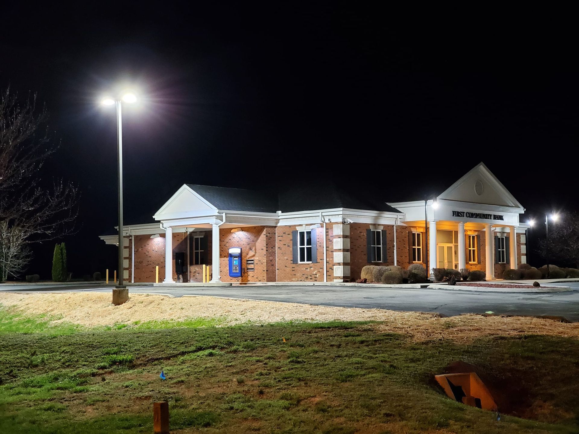 A red brick bank building with white columns at night, illuminated by bright parking lot lights.