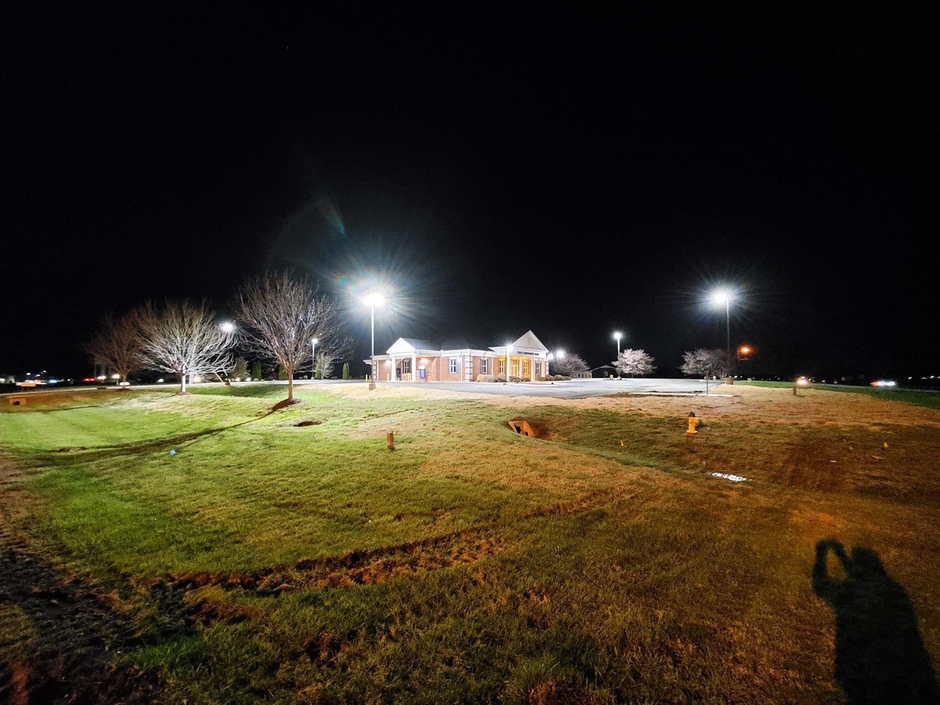 A brightly lit building stands at night on a grassy lot, surrounded by tall light poles under a dark, clear sky.