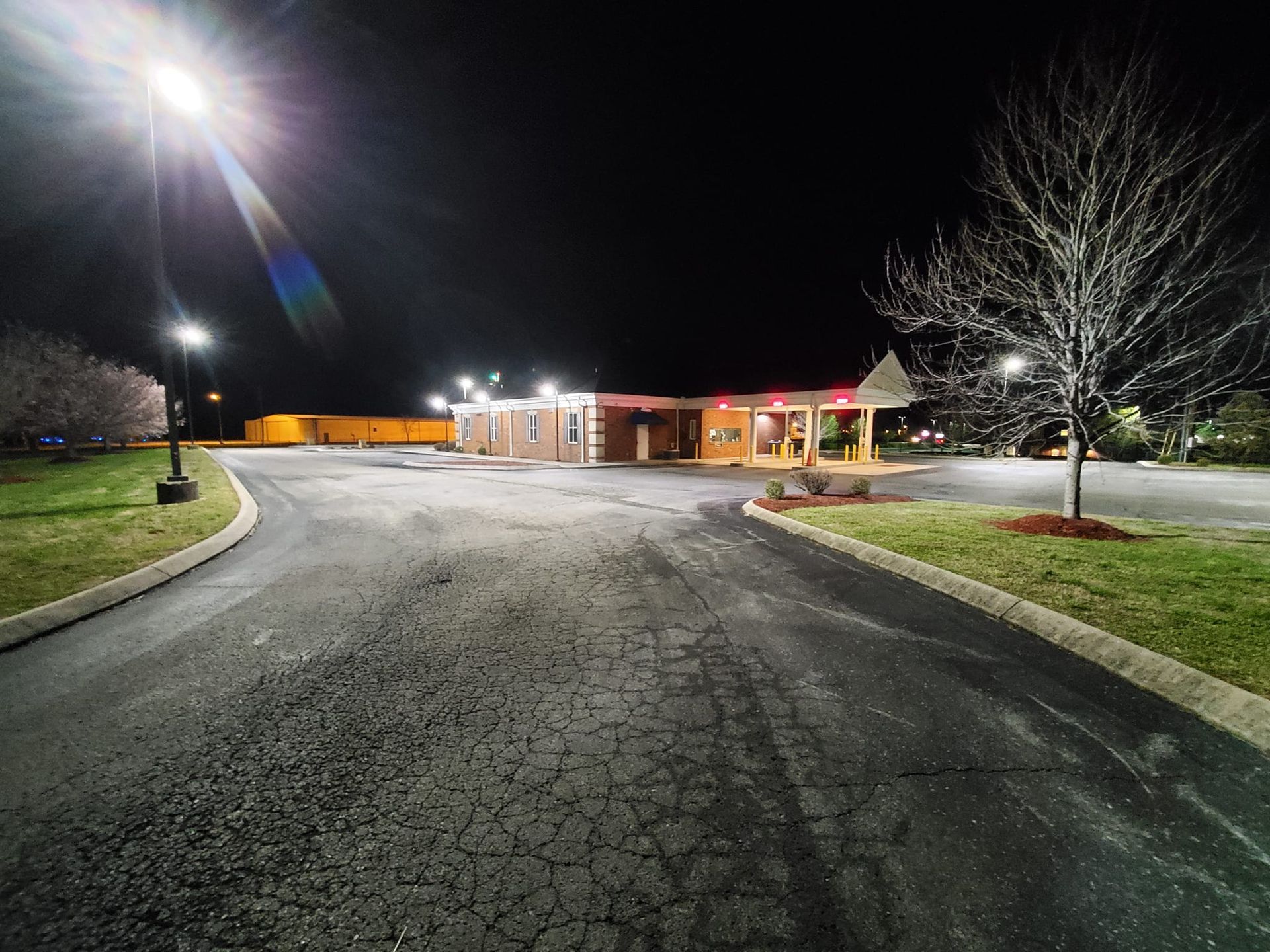 A dimly lit parking lot at night in front of a small, illuminated building with a dark sky and a leafless tree.