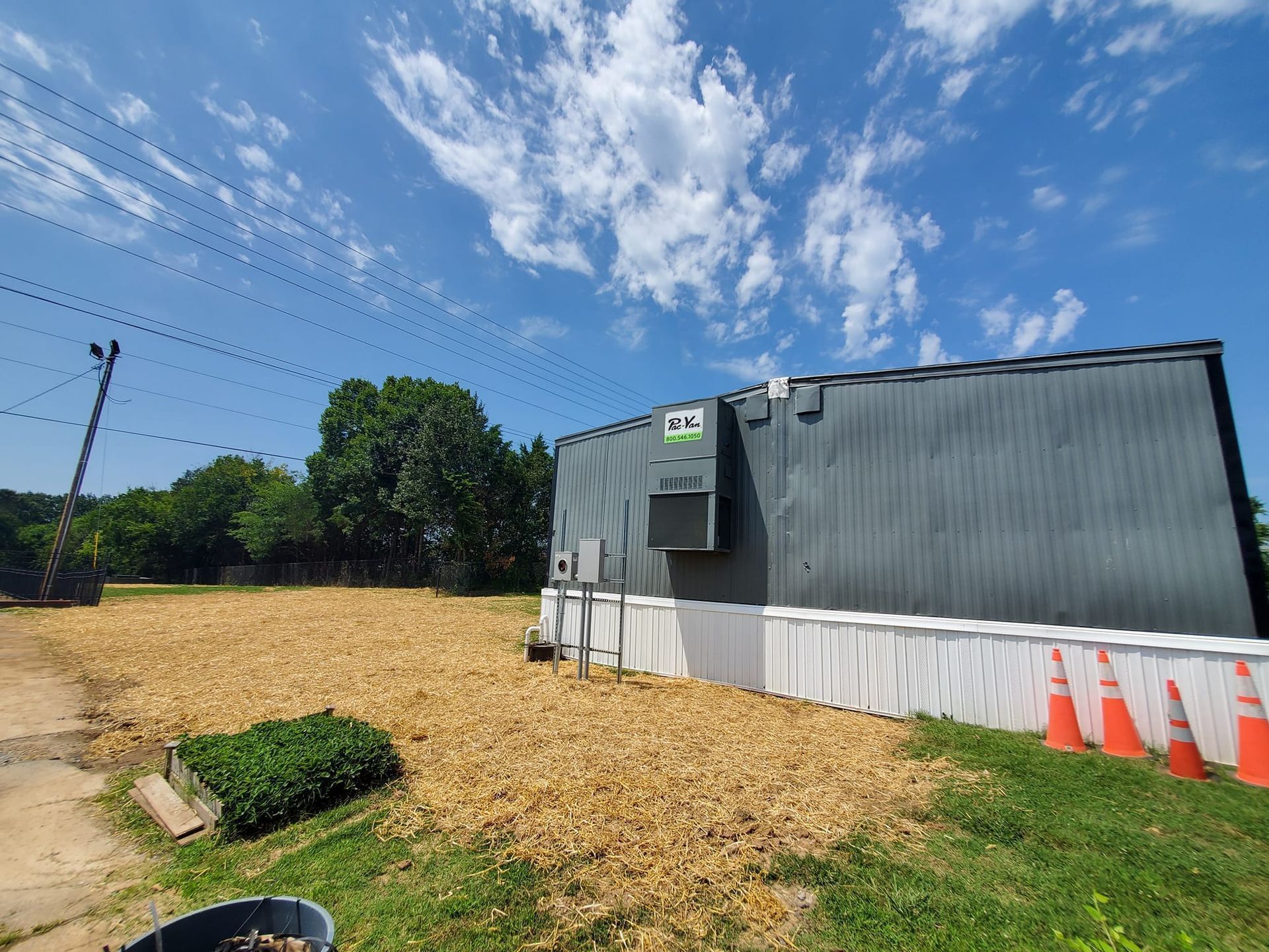 A dark grey storage building with white skirting sits on a lot covered in wood chips under a bright blue sky.
