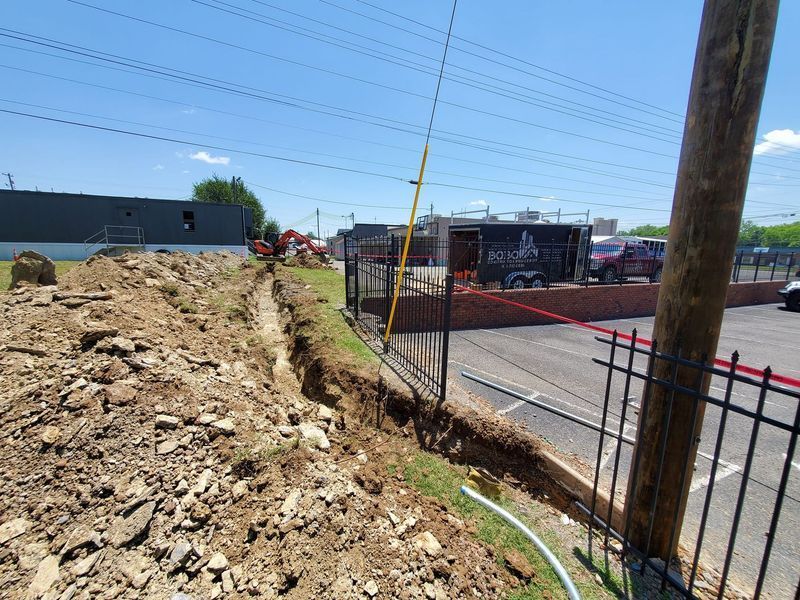 A construction trench runs alongside a metal fence and parking lot, with a building and equipment in the background.