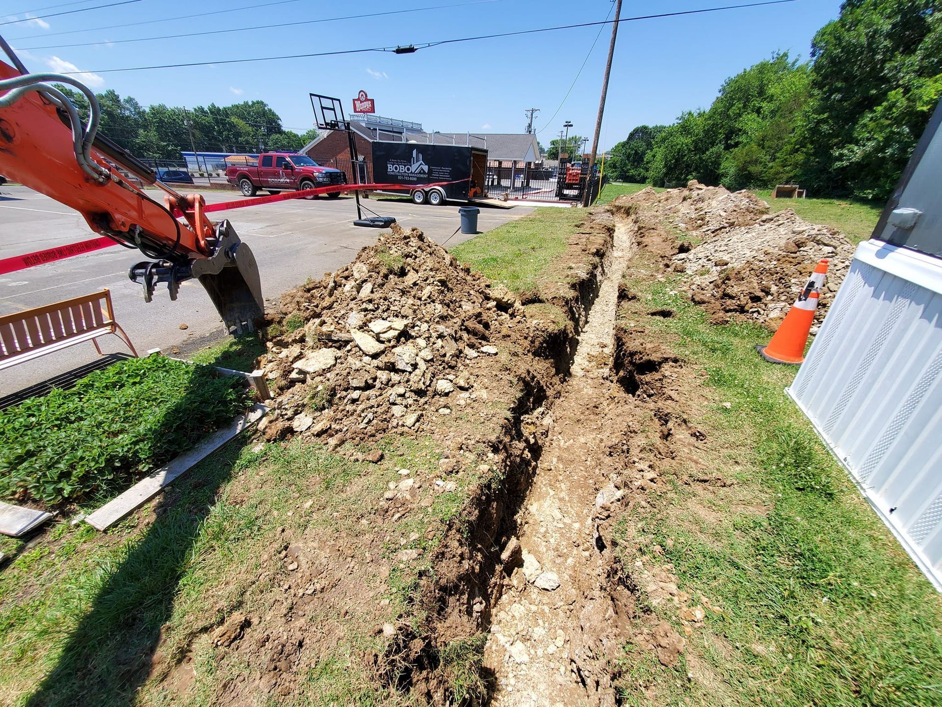 An excavator arm extends over a grassy area where a long, narrow trench has been dug beside a building.