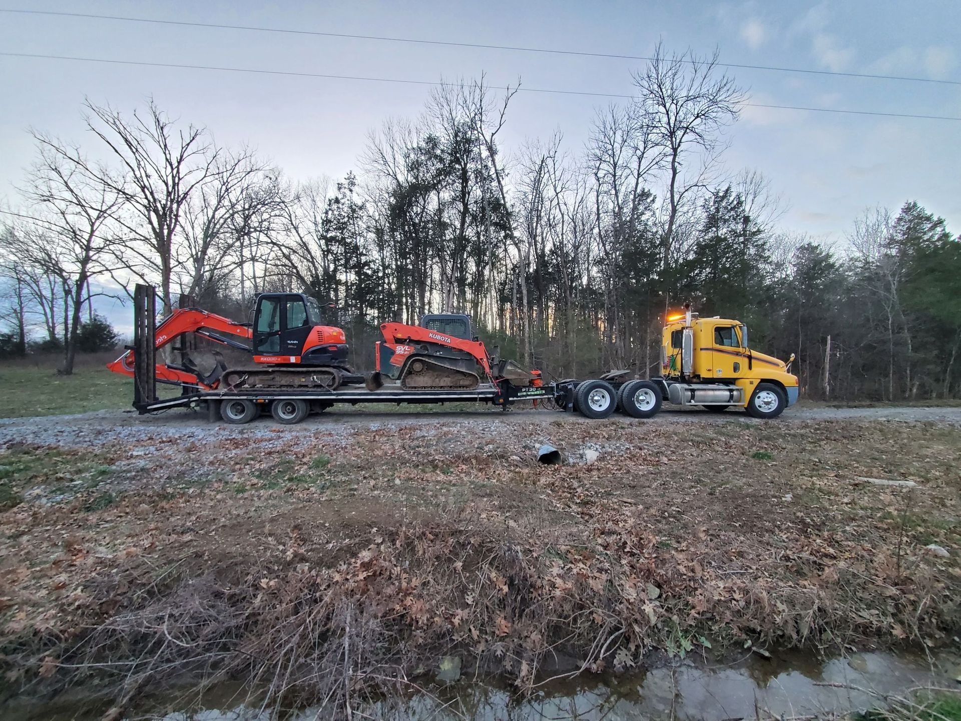 A yellow semi-truck pulling a flatbed trailer loaded with orange construction equipment on a rural, leaf-covered road.