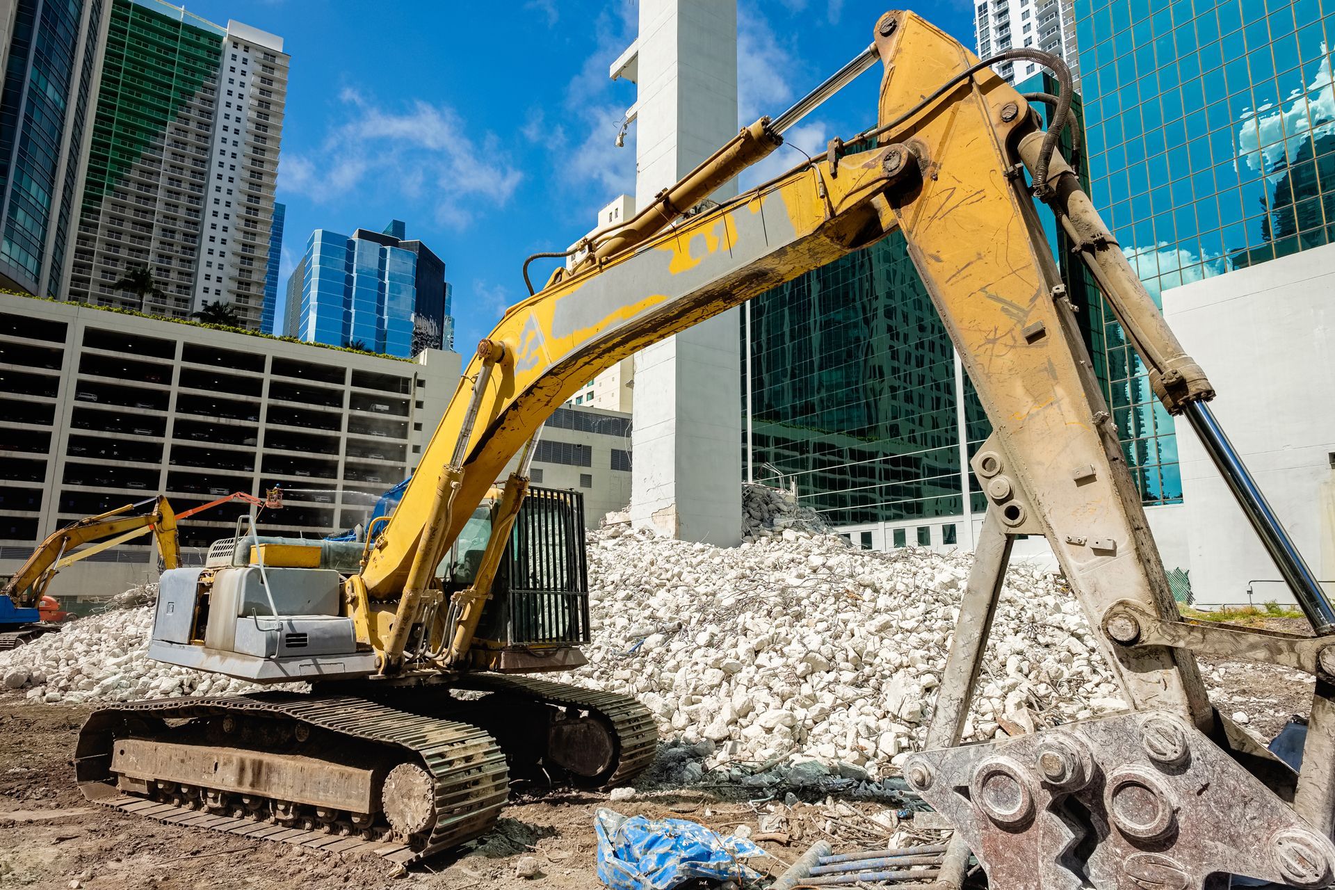A yellow excavator stands at a construction site with piles of debris and modern office buildings in the background.