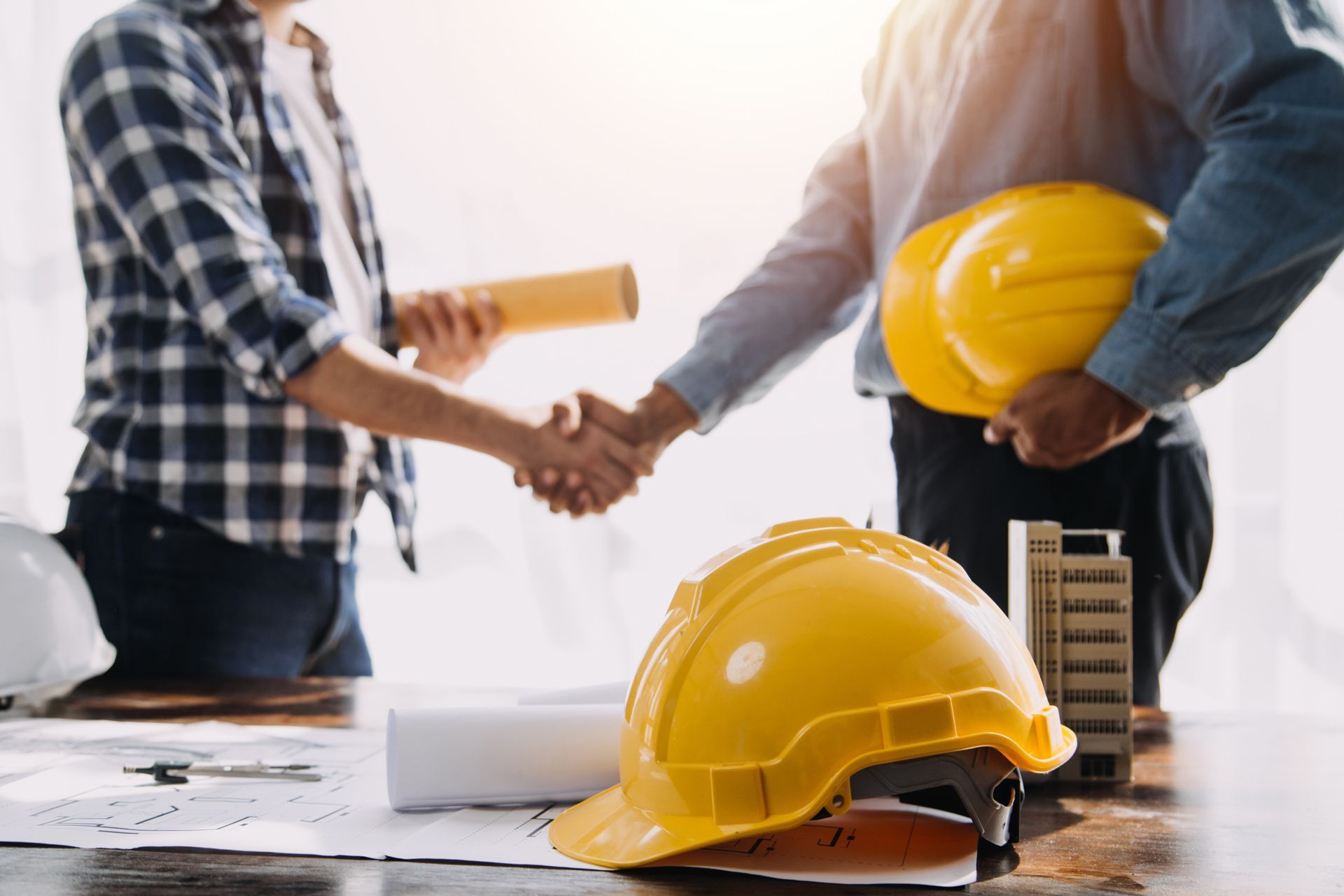 Two professionals in work clothes shake hands over building plans, with yellow hard hats nearby on a wooden desk.
