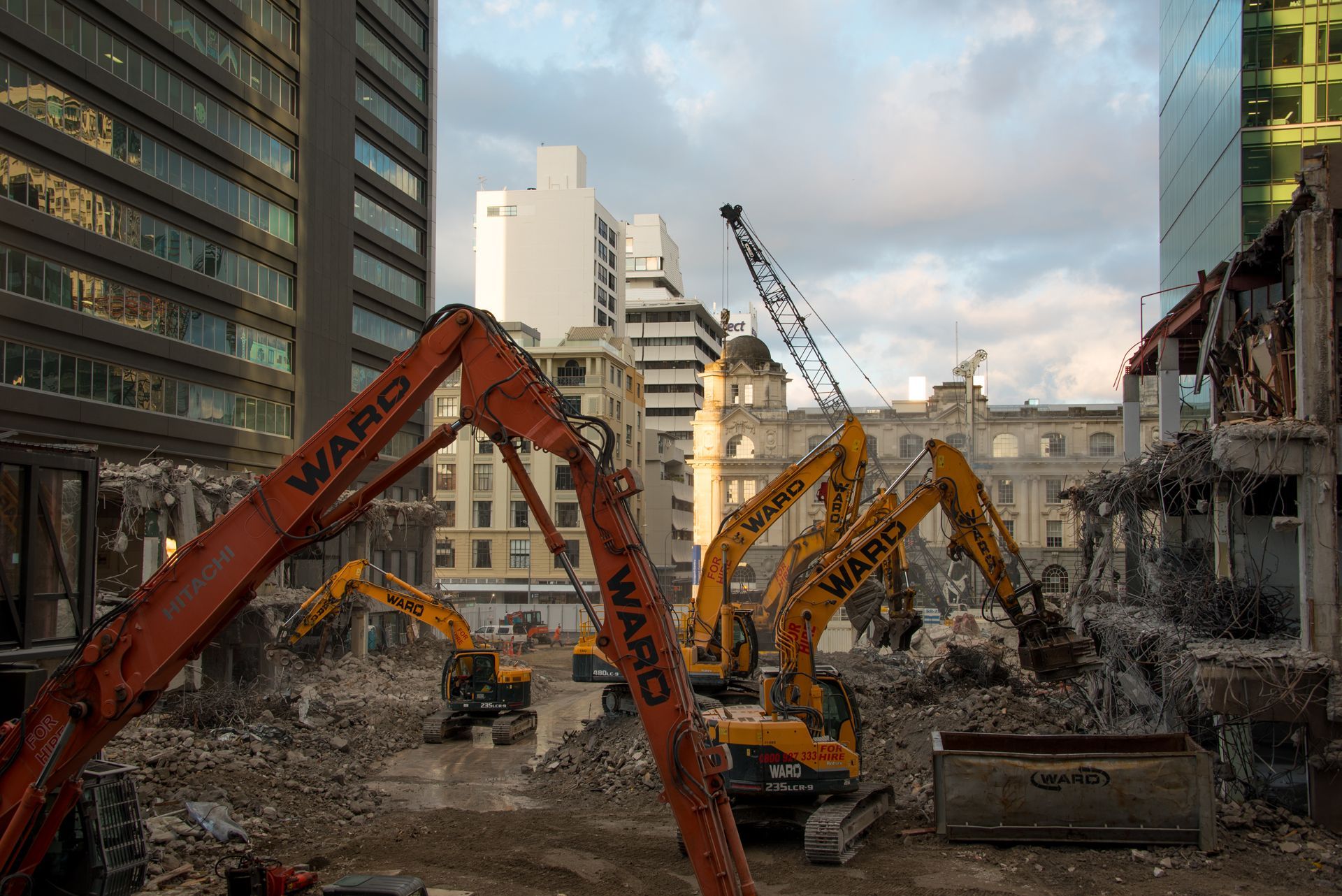 Multiple orange and yellow excavators work on a debris-filled urban construction site surrounded by city buildings.