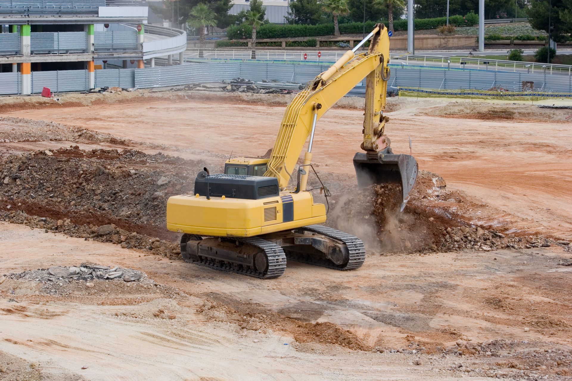 A yellow tracked excavator digs into a large, dusty construction site.