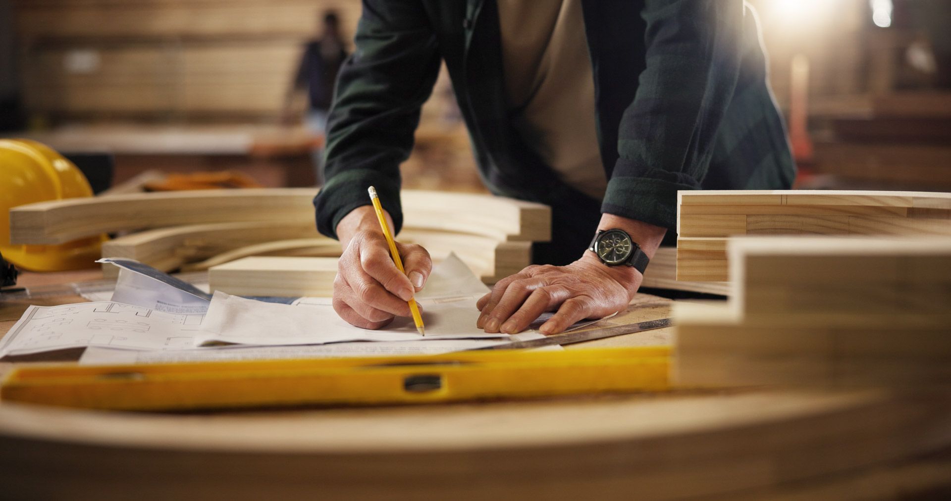 Carpenter using a pencil to draw on blueprints at a workbench with wood pieces and a level in a workshop.