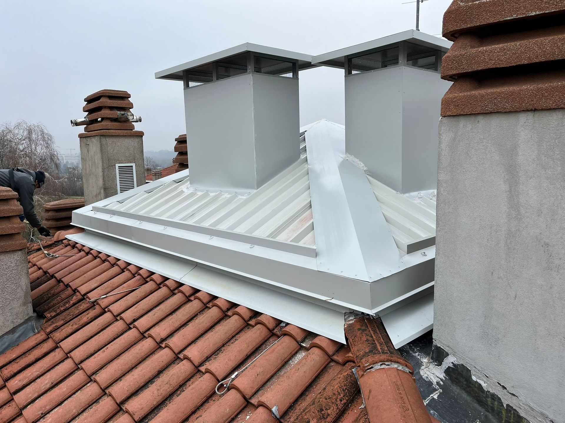 Tetto con tegole rosse, sezione del tetto in metallo bianco e due camini quadrati e bianchi contro un cielo nuvoloso.