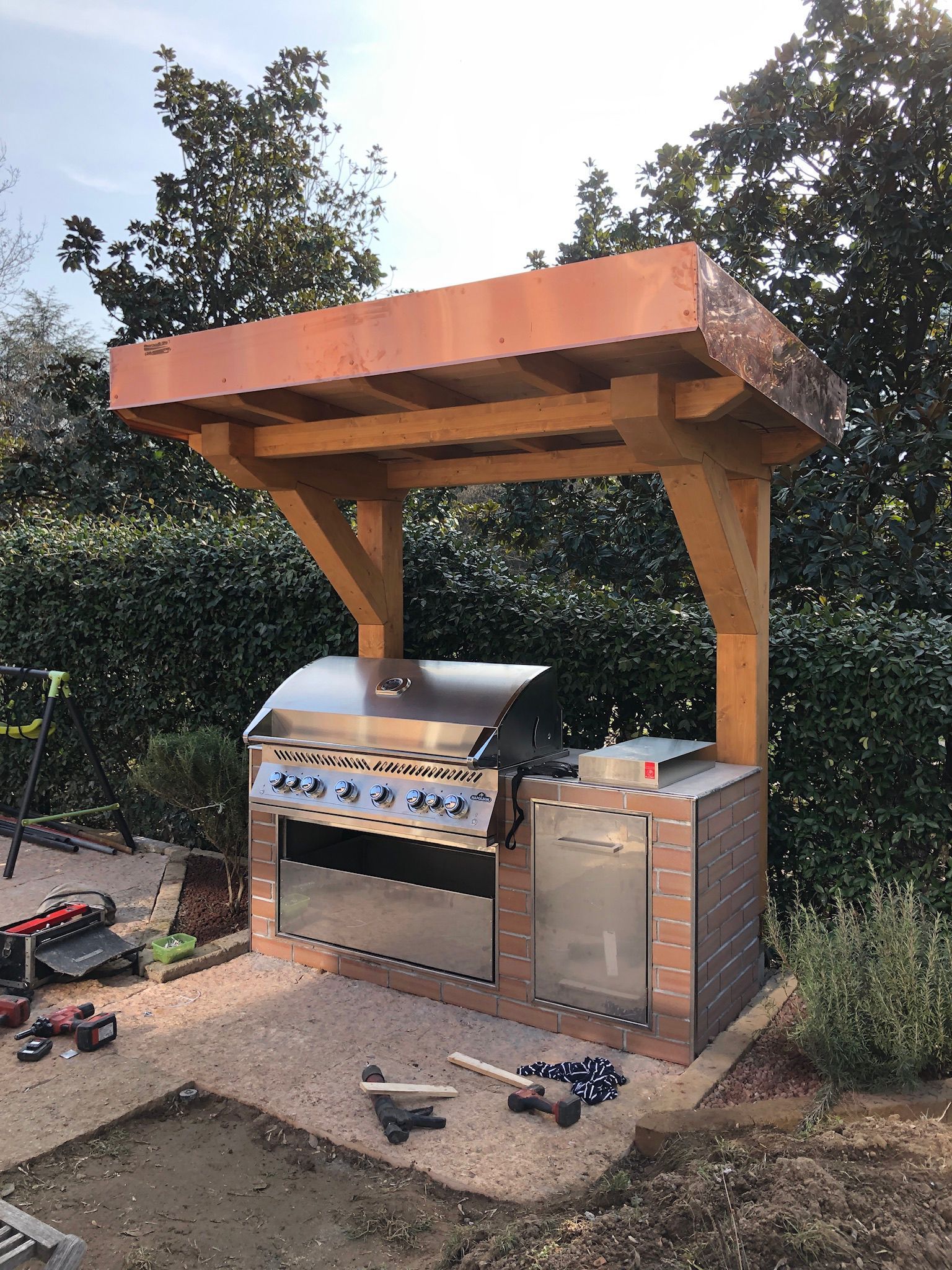 Cucina da esterno con griglia in acciaio inox e base in mattoni, sotto una tettoia in legno.
