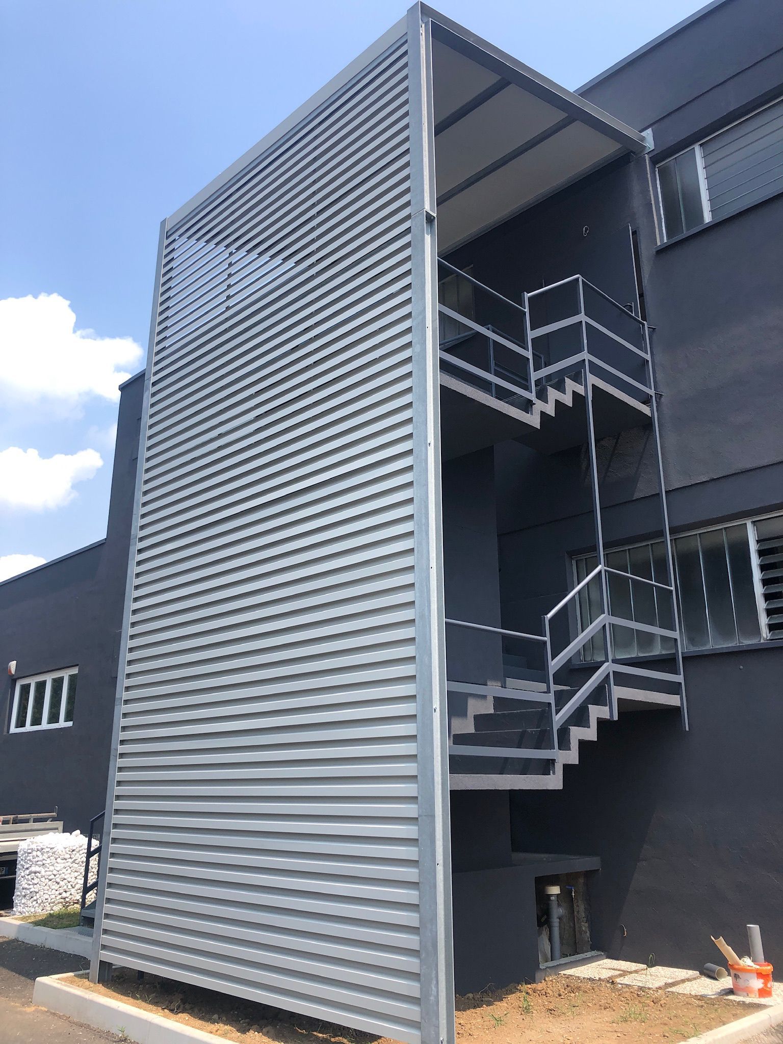 Scala esterna in metallo dietro una parete grigia perforata fissata a un edificio scuro.