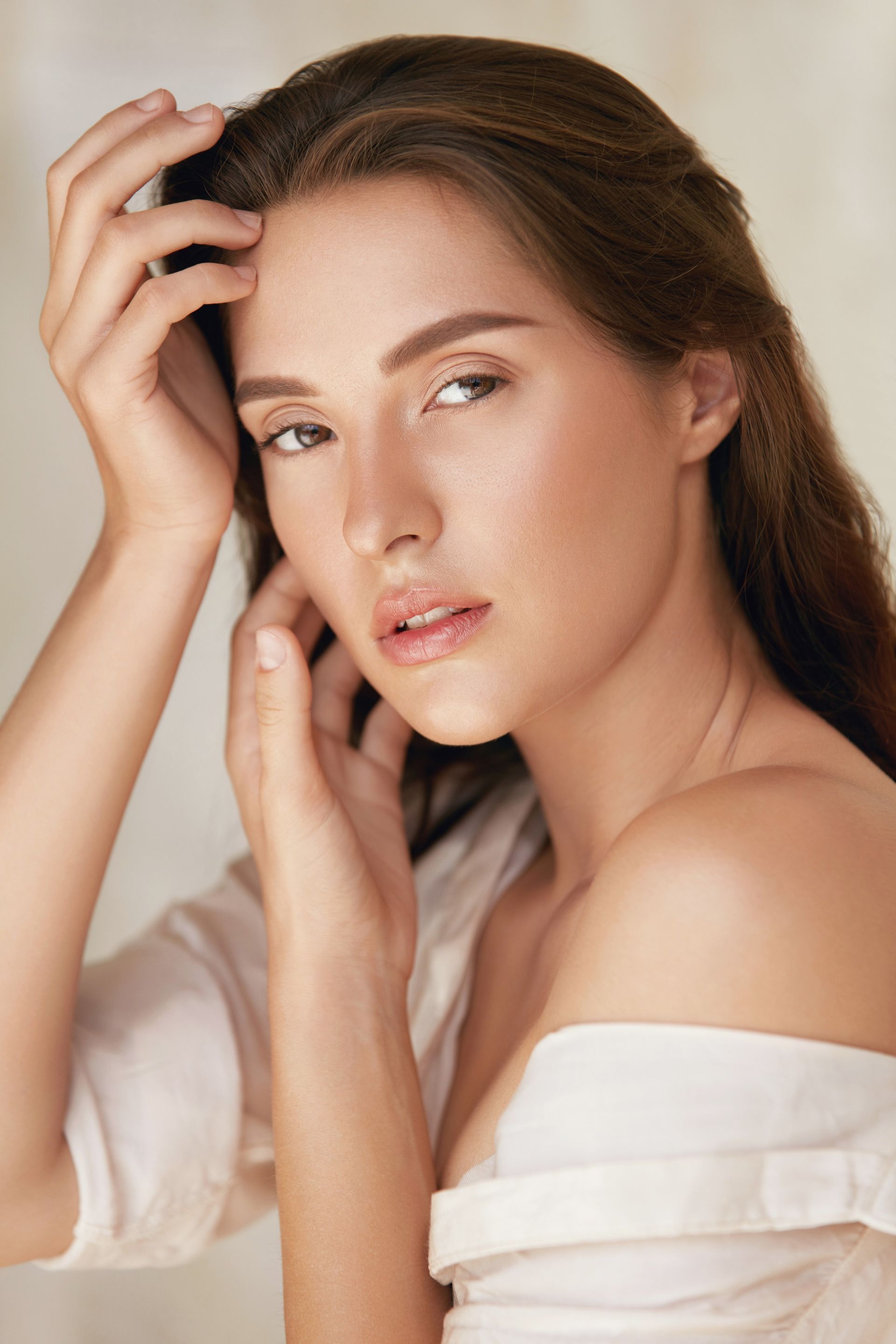 A person with dark hair looks towards the camera, hands posed near their face, wearing a white shirt against a light wall.