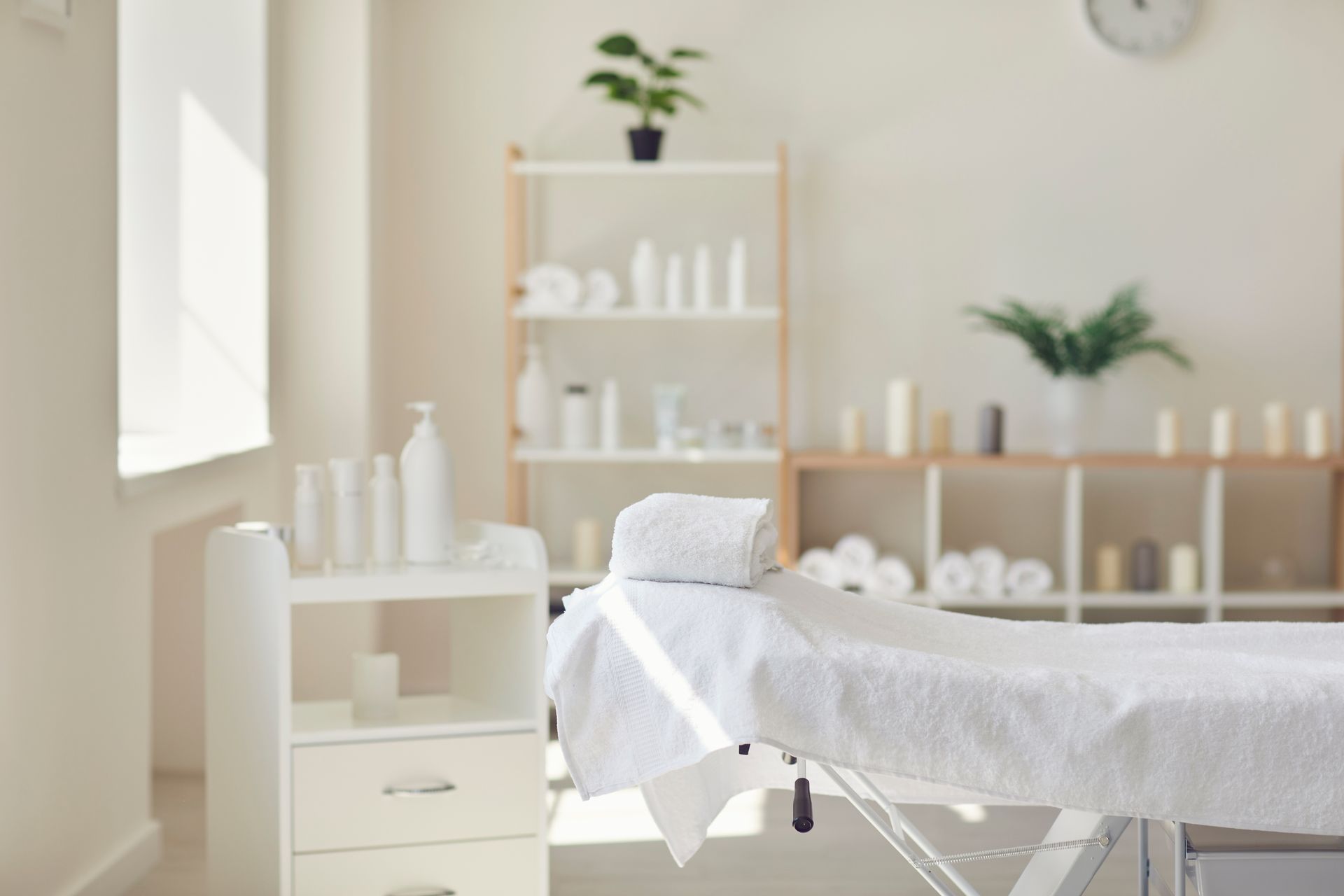 A clean, modern spa treatment room with a massage table, white linens, shelves with products, and green plants.