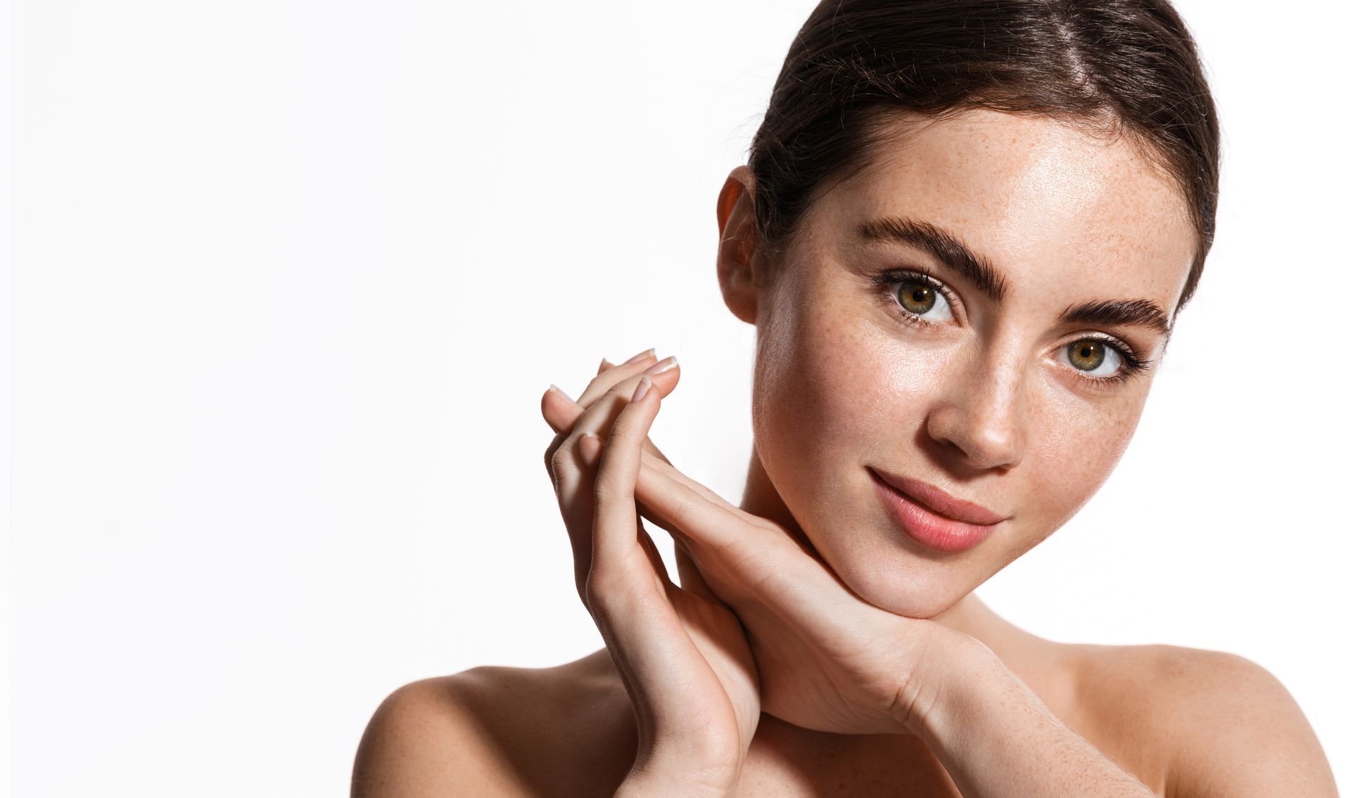 Close-up portrait of a person with freckles resting their chin gently on their hands against a plain white background.