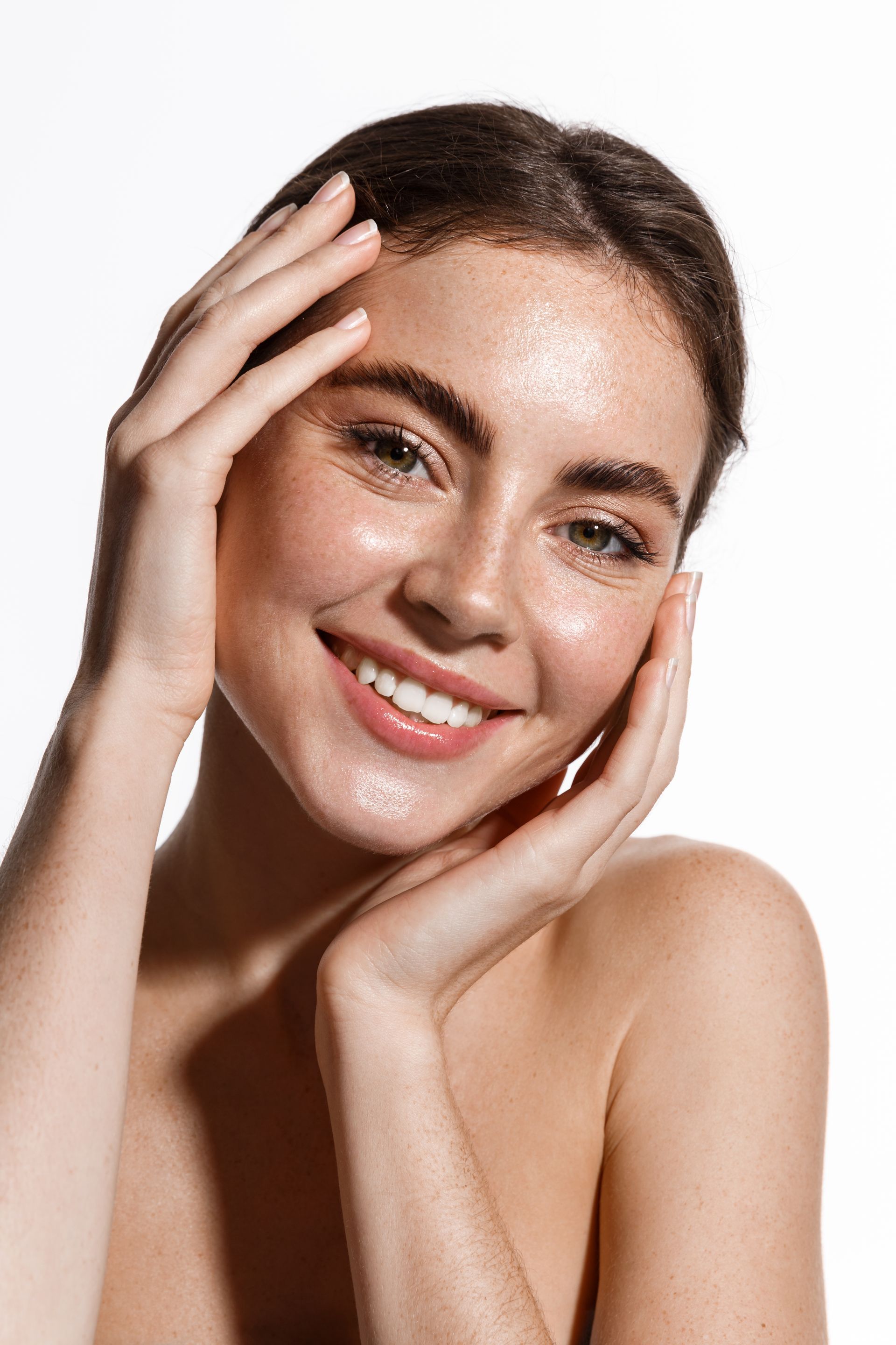 A smiling person with freckled skin and a dewy complexion rests their hands against their cheeks, facing the camera.