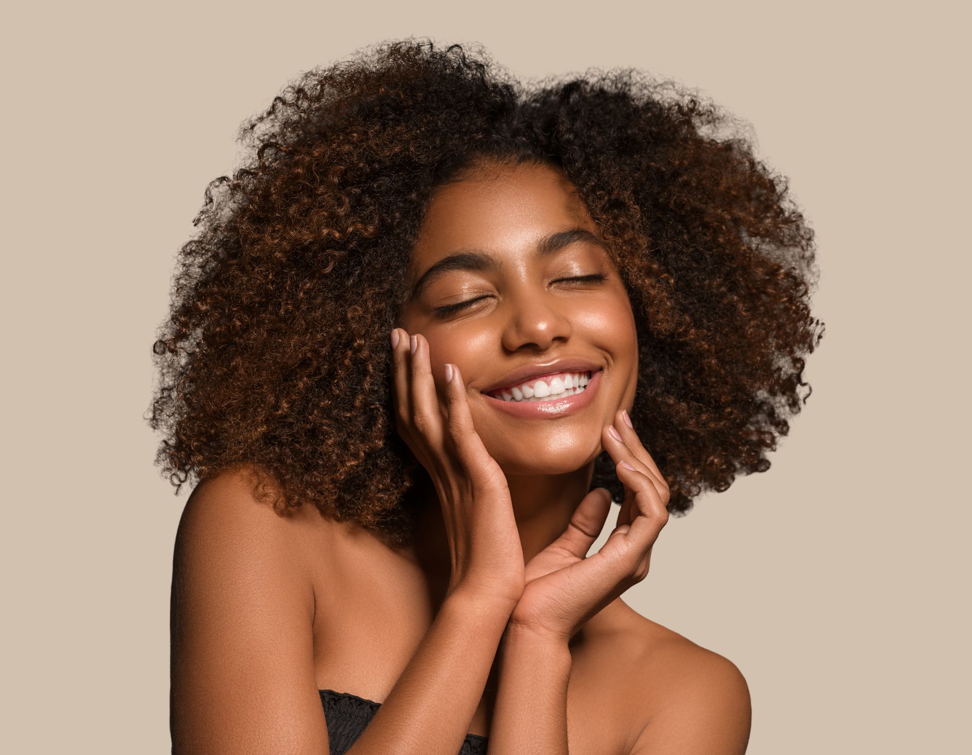 A woman with curly hair is smiling and touching her face with her hands.