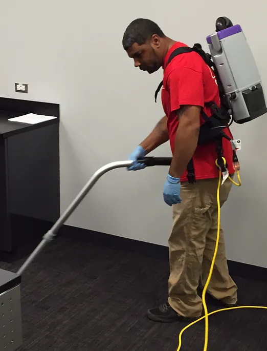 Person in red shirt and tan pants vacuums carpet with backpack vacuum in an office setting.