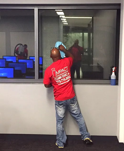 Man in red shirt cleaning a window with a blue cloth and spray bottle. Office setting.