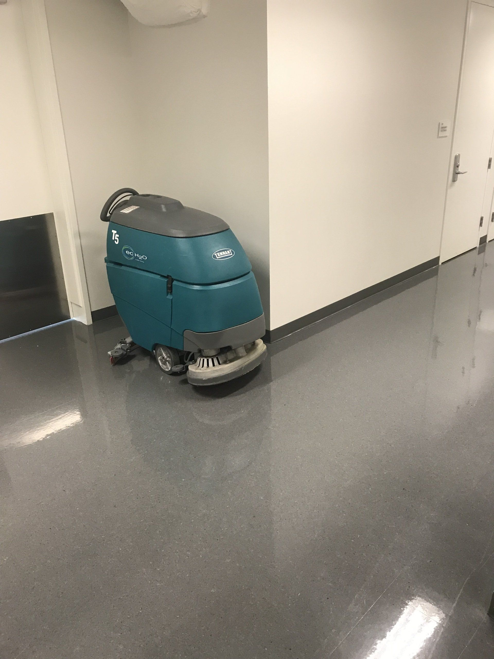 A teal floor cleaning machine is parked in a hallway with shiny gray floors and white walls.