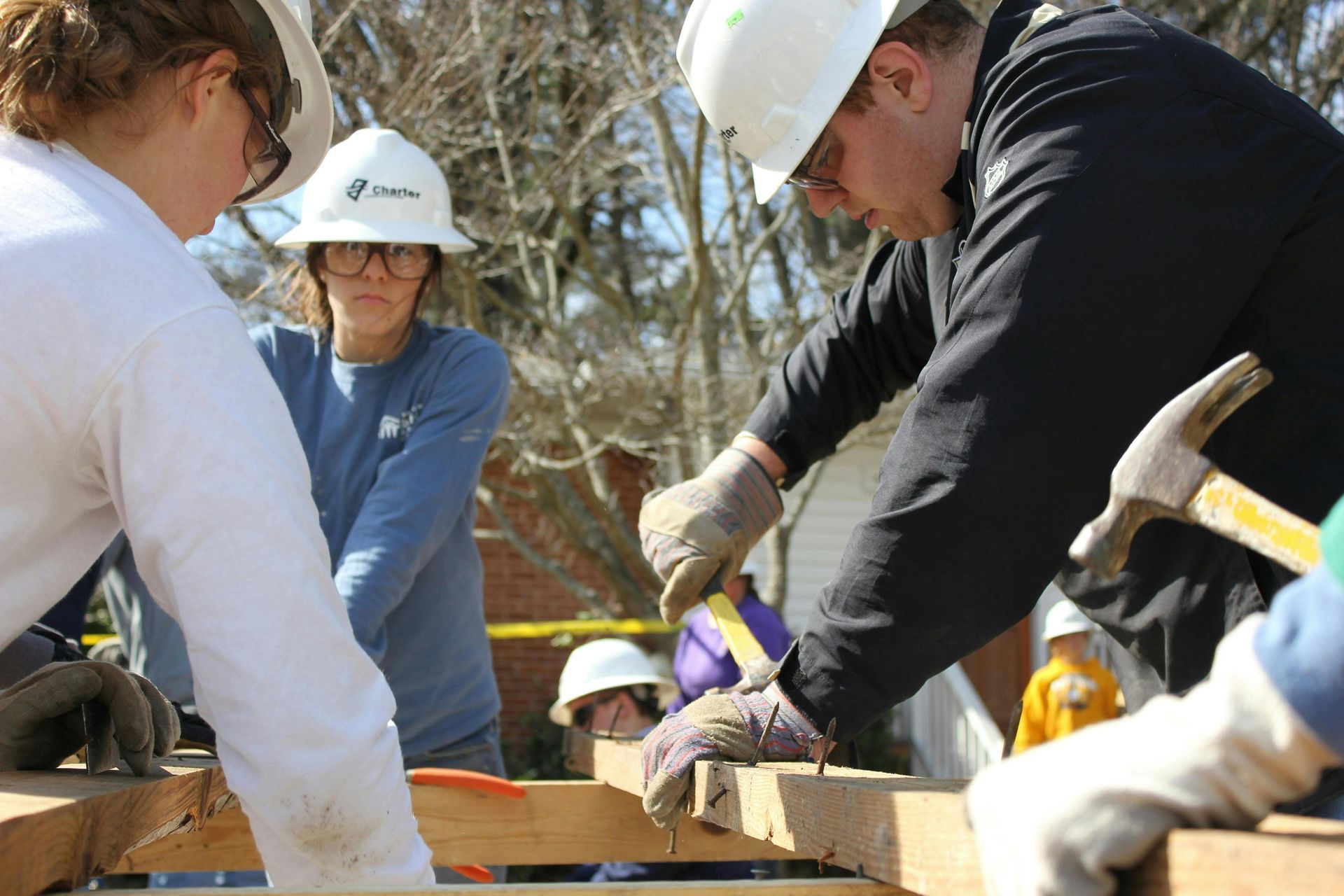 People in hard hats building with wood, using hammers. Outdoors, sunny day.