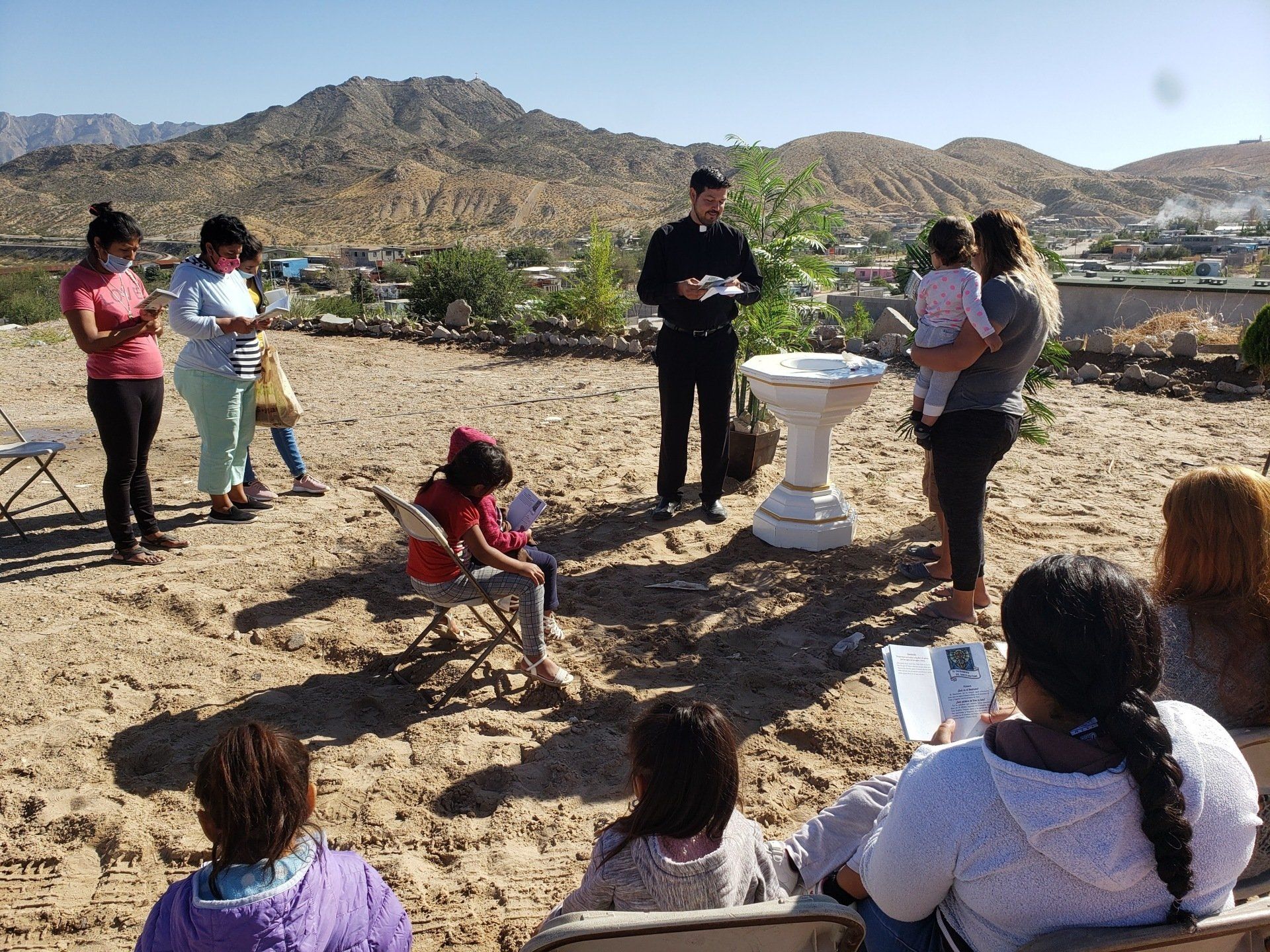 A group of people are sitting in chairs in the sand in front of a man holding a baby.