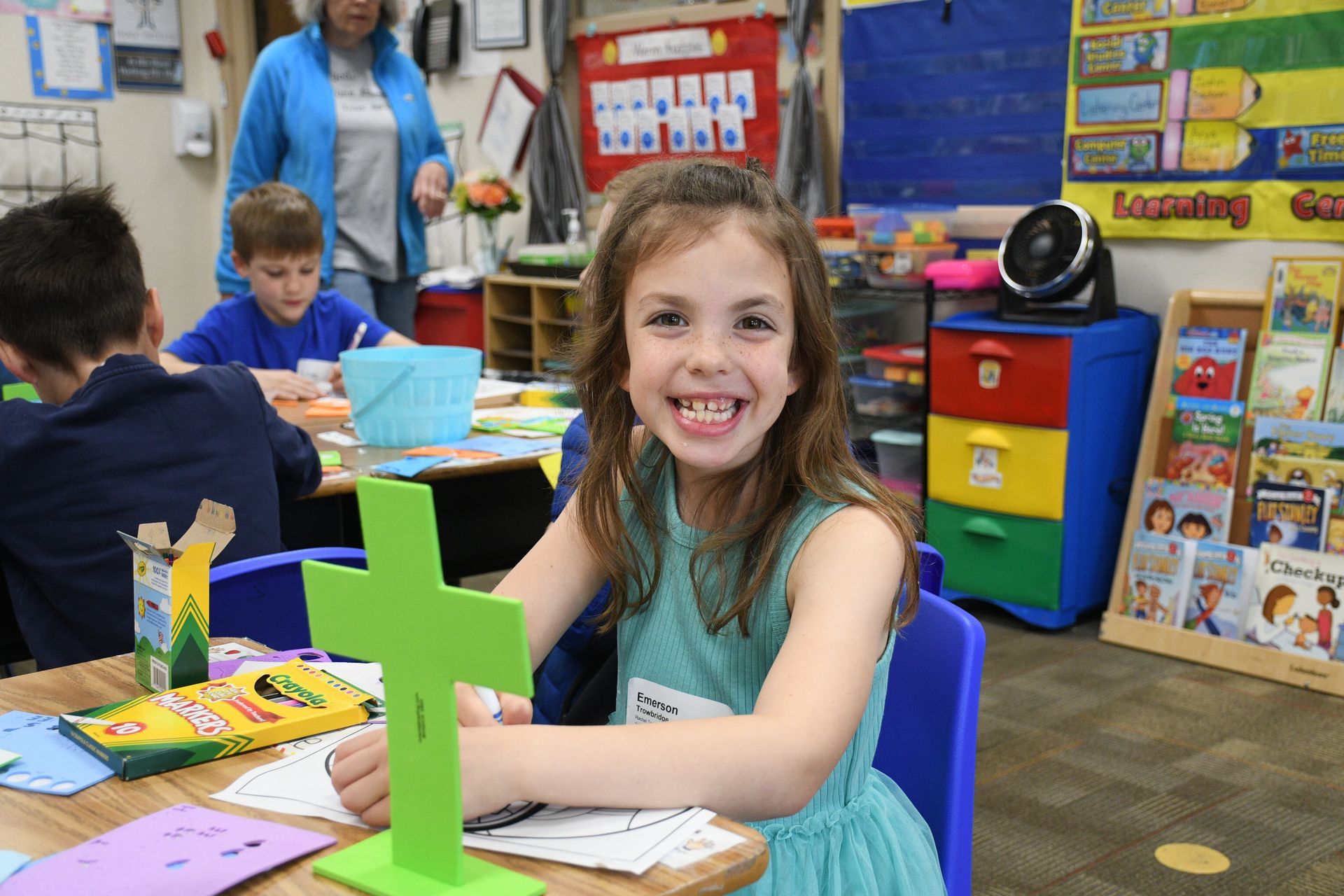 A little girl is sitting at a table in a classroom holding a green cross.