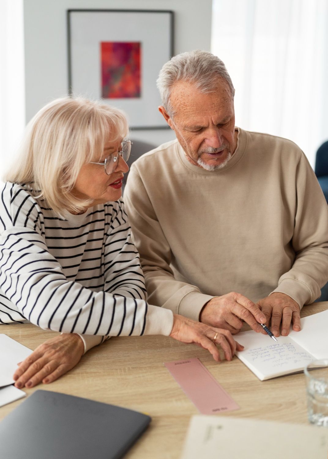 Older couple reviewing documents at a table. Woman points; man looks intently. Neutral beige and white tones.
