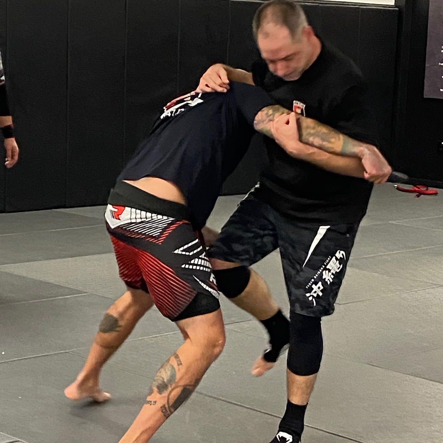 Two men are wrestling in a gym and one has a tattoo on his arm