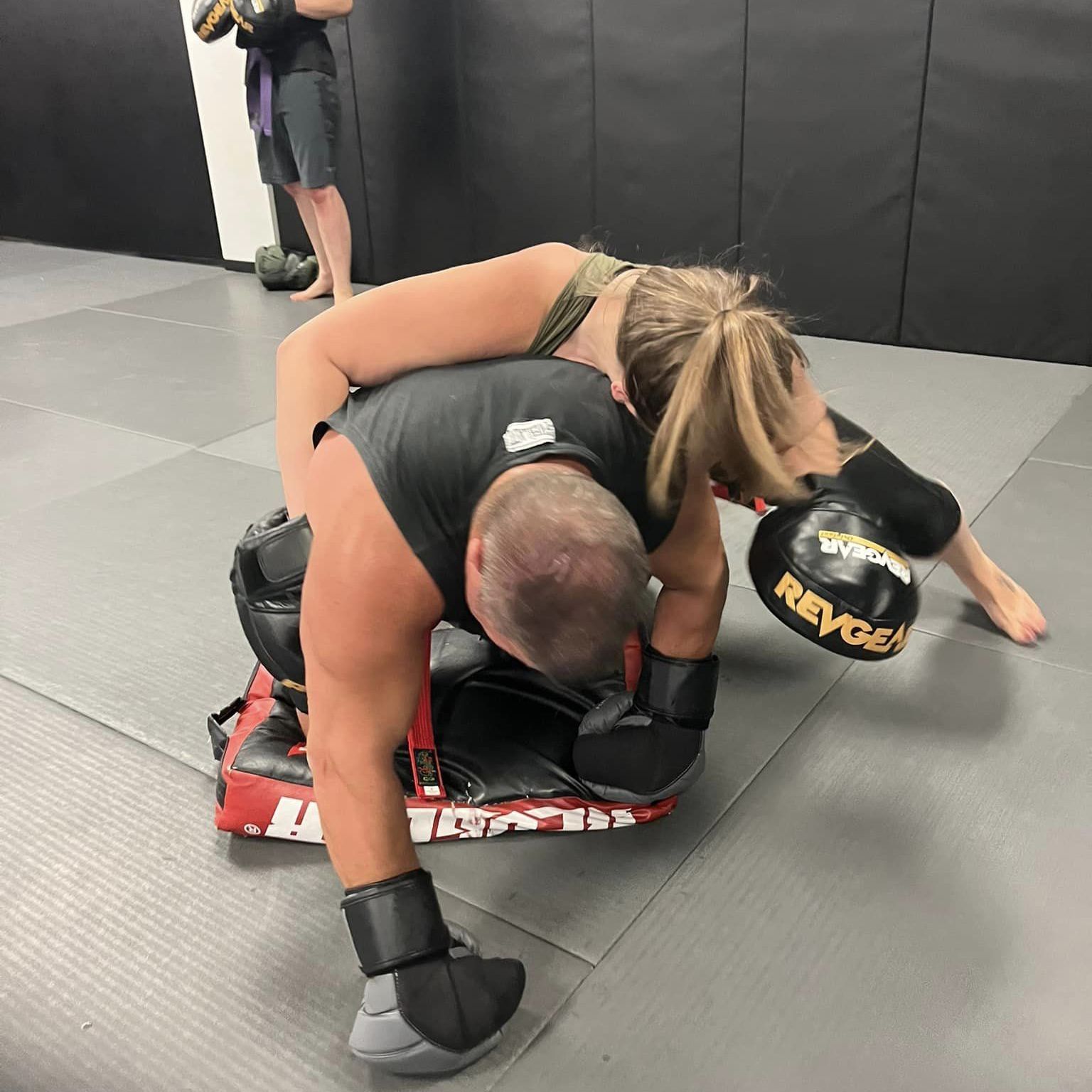 A man and a woman are wrestling in a gym and the woman is wearing a pair of reyes boxing gloves