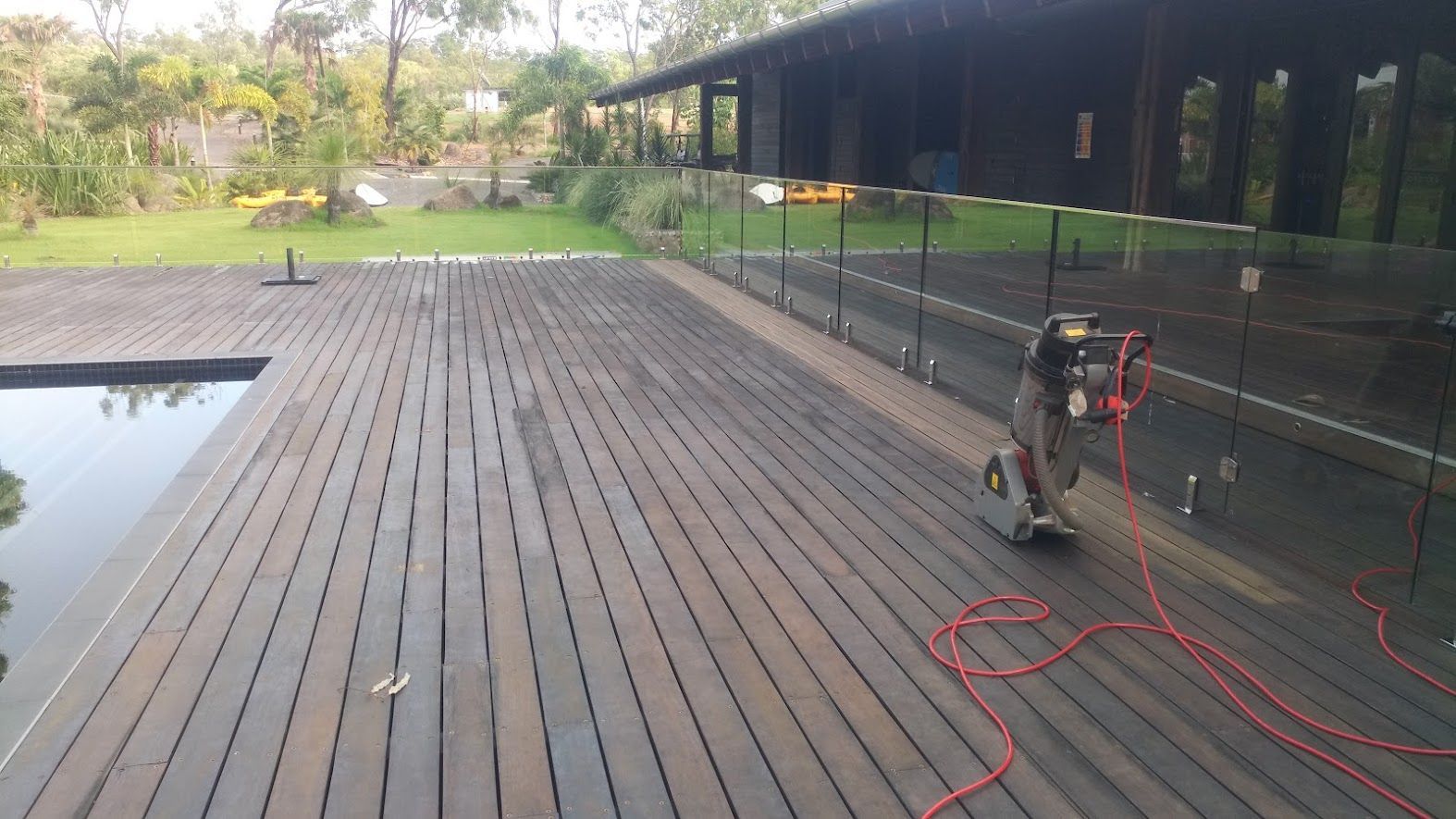 Wooden deck being cleaned with a pressure washer, next to a pool and house with glass railing.