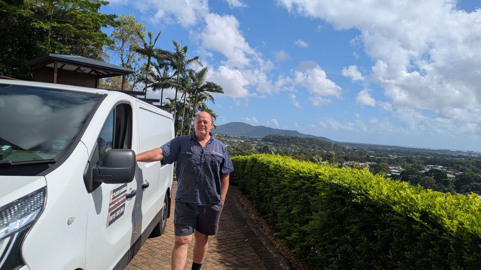 Man standing beside white van on hill overlooking a town under a blue, cloudy sky.