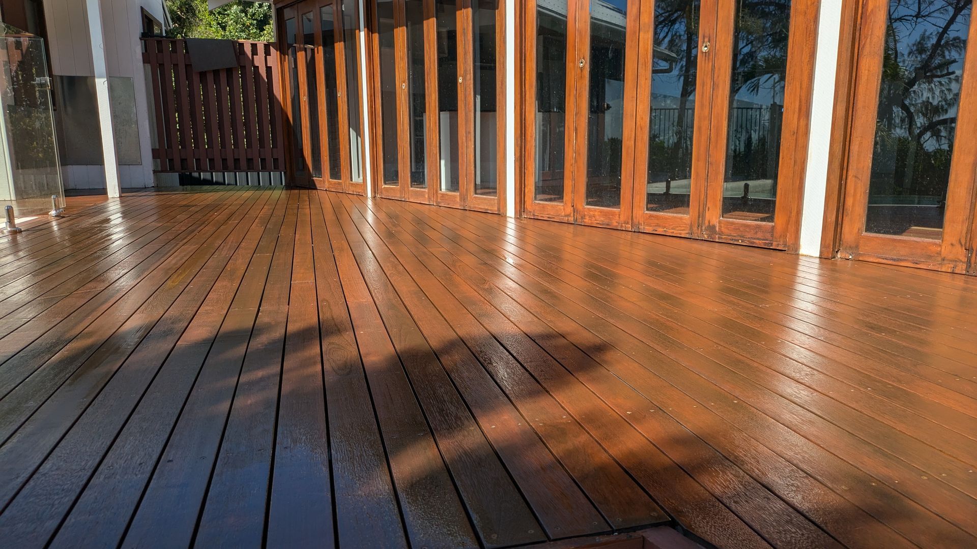 Wooden deck with a glossy finish, reflecting sunlight. Doors and a railing are visible in the background.