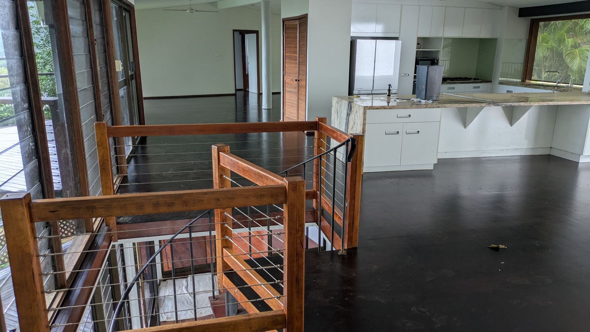 Interior of an empty home with dark floors, wooden railing, and a kitchen area.
