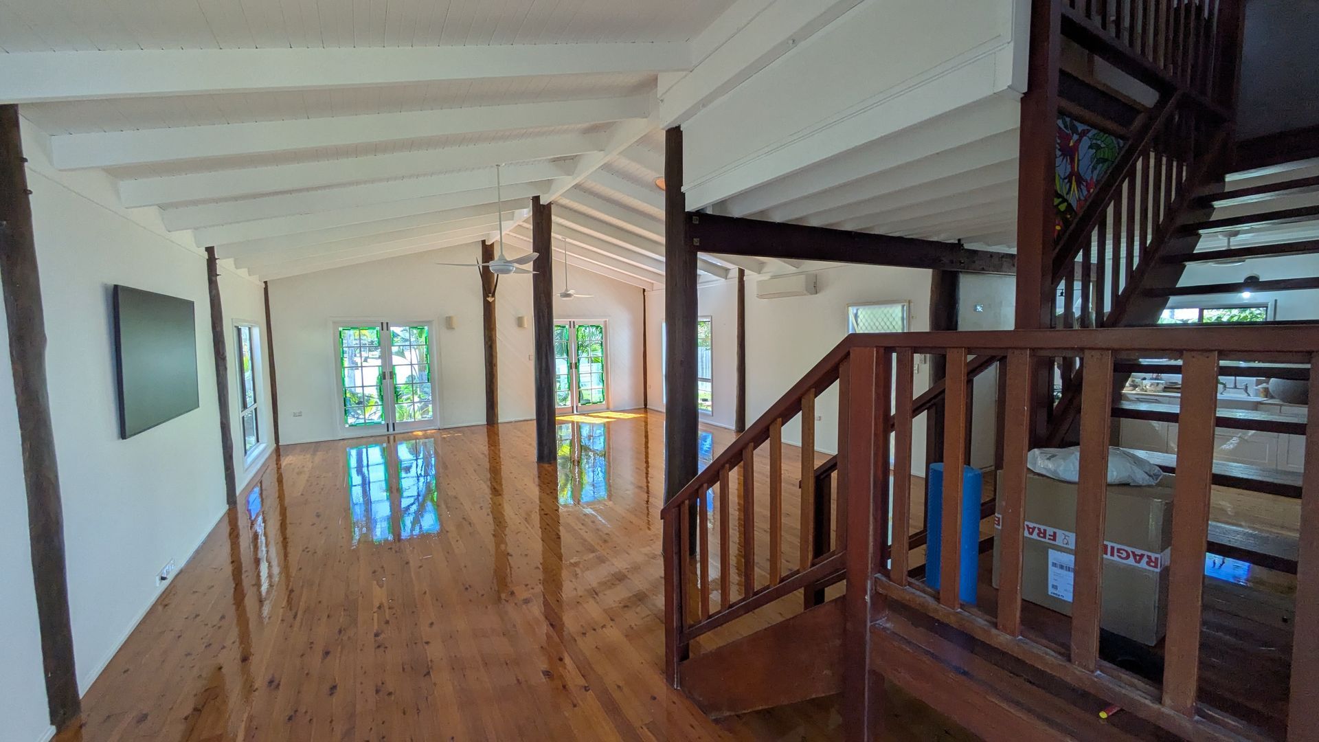 Spacious living area with wooden floor, white ceiling, and a staircase. Sunlight streams in from glass doors.